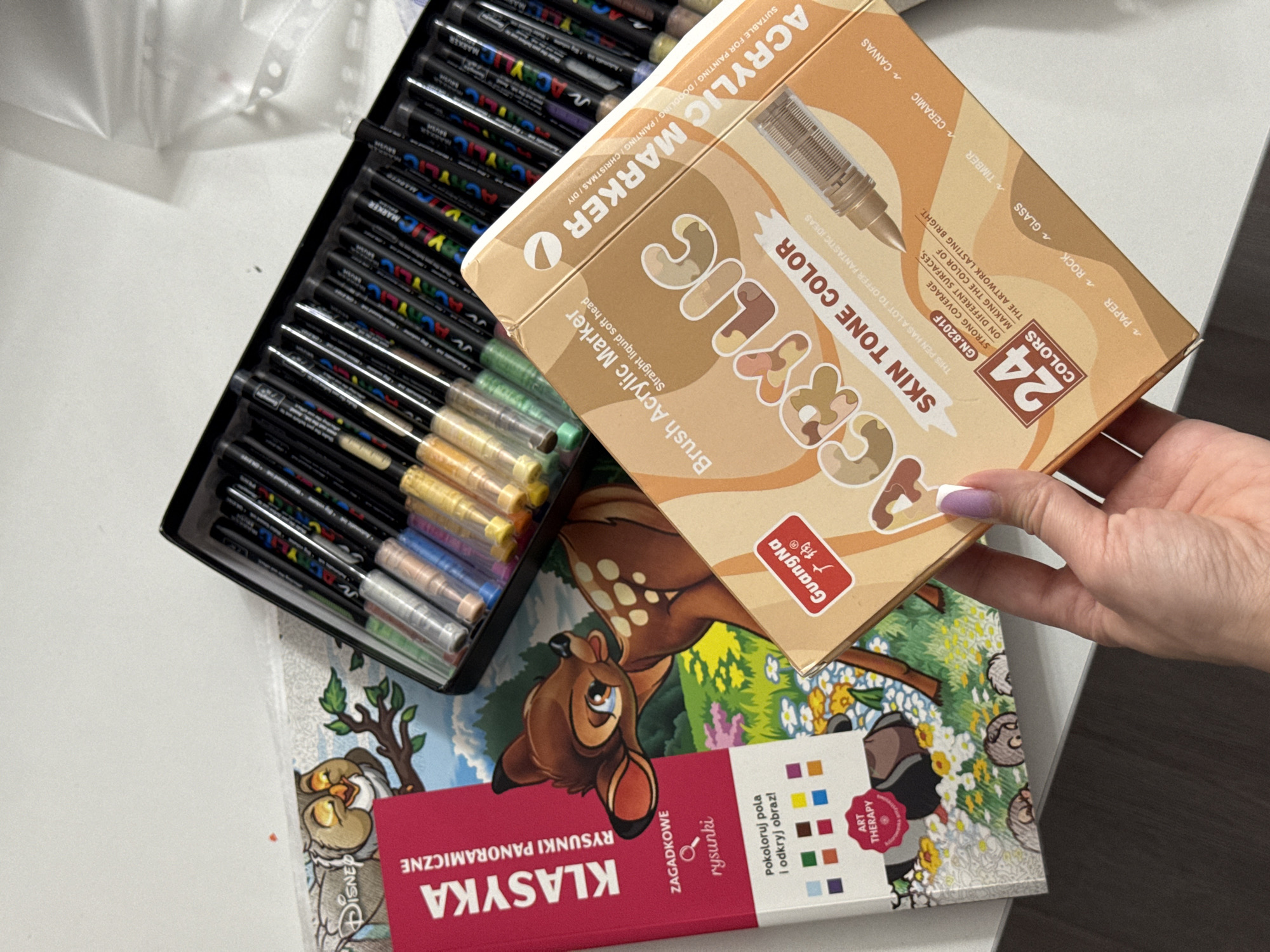 The image shows a flat lay of art supplies on a white surface. In the foreground, a hand with purple-tipped nails holds a box of "ACRYLIC MARKER SKIN TONE COLOR" pens. The box is orange and tan and displays the brand "GuangNa." Behind the box of markers, a coloring book featuring Bambi from Disney is visible. The coloring book is pink and has Polish text, including "KLASYKA" and "RYSUNKI PANORAMICZNE." A metal case filled with various colored acrylic markers is positioned in the upper right portion of the image. The overall scene suggests a creative setup or the recent acquisition of art materials. There is no visible indication of the location being in the city of Łódź, Poland, or any other specific city.