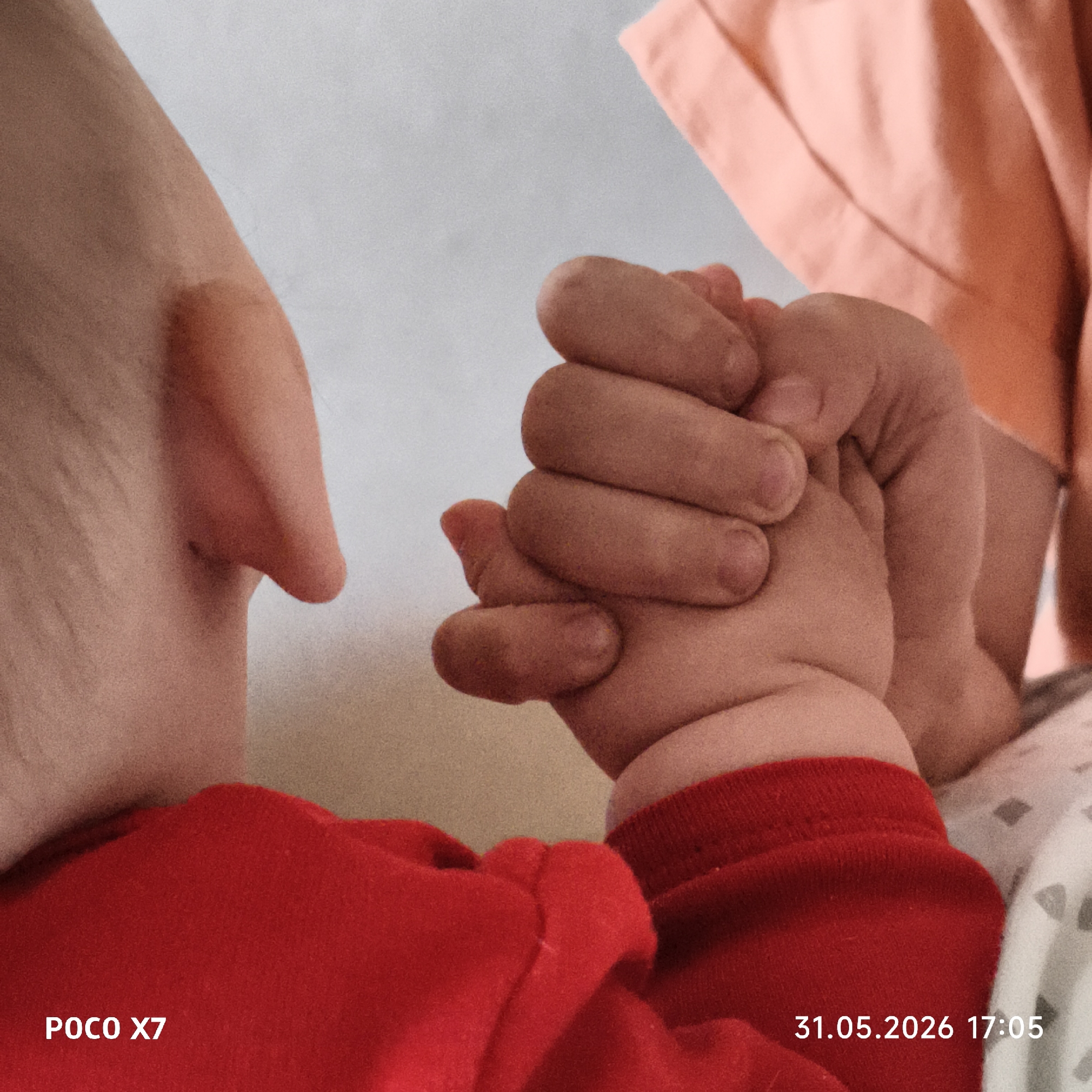 A close-up shot shows an adult's hand intertwined with a baby's hand. The baby wears a red garment. The adult appears to be wearing a peach-colored garment. The bottom left corner contains the text "POCO X7." The bottom right corner shows "31.05.2026 17:05." The location in the city of Unknown, Unknown is not determined.