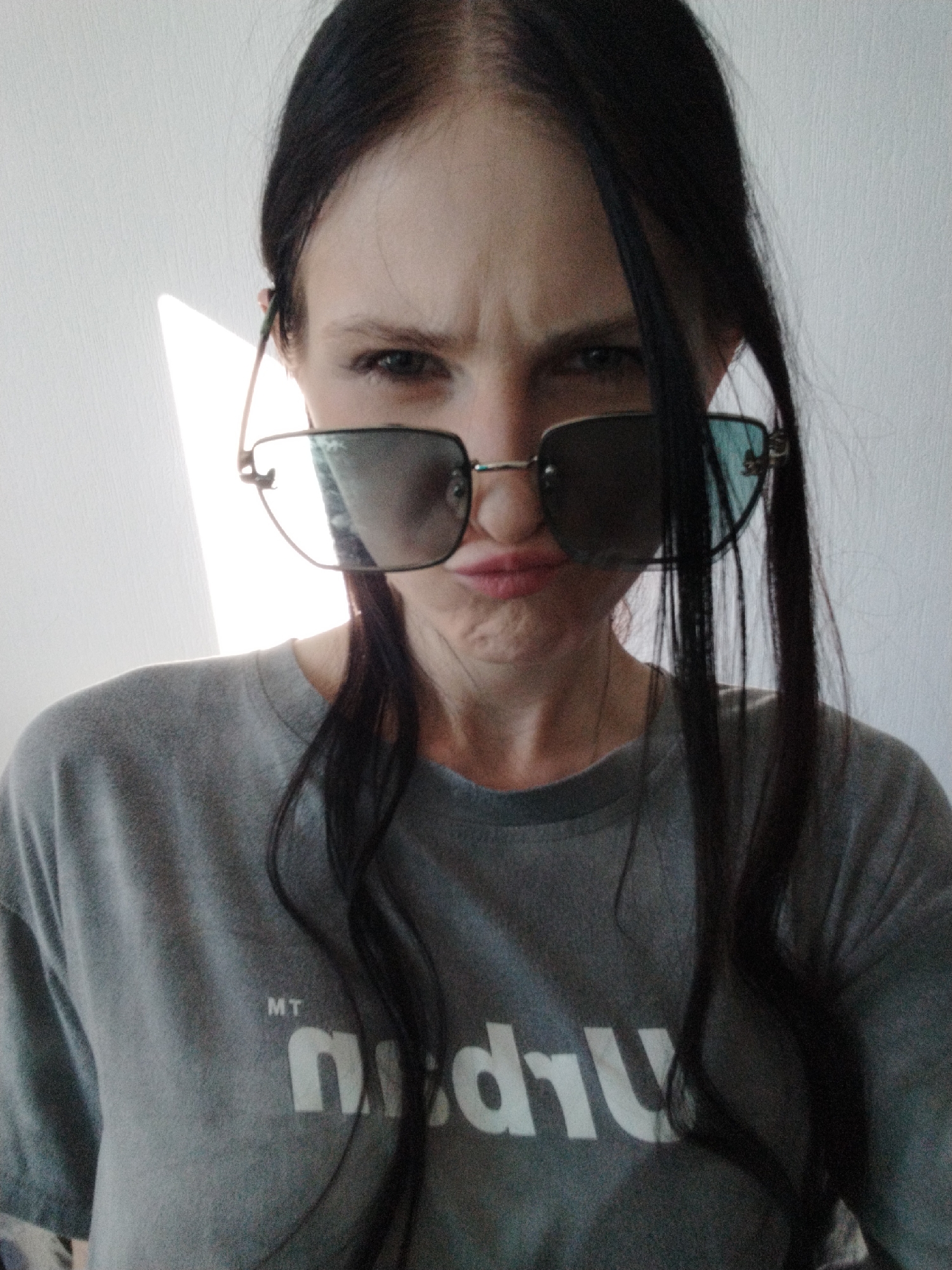 This media file is a close-up, waist-up, selfie-style photograph of a person wearing sunglasses and a grey t-shirt. The person has dark hair that is styled in loose waves and partially obscures their face. They are looking directly at the camera with a slightly furrowed brow and a playful, perhaps slightly annoyed, expression, with their lips pursed. The sunglasses are large and square-framed with tinted lenses. The grey t-shirt has a graphic logo on the front. Visible text on the t-shirt includes "MT" in small letters above a larger, stylized graphic that appears to read "nuDU". The background is a plain, light-colored wall, with a sliver of bright light entering from the left side, suggesting either a window or a light source. The lighting is somewhat harsh, creating shadows and highlights on the person's face and t-shirt. The overall impression is casual and somewhat humorous, possibly taken indoors during the day. The lack of discernible background elements makes the exact location and time of day unclear.