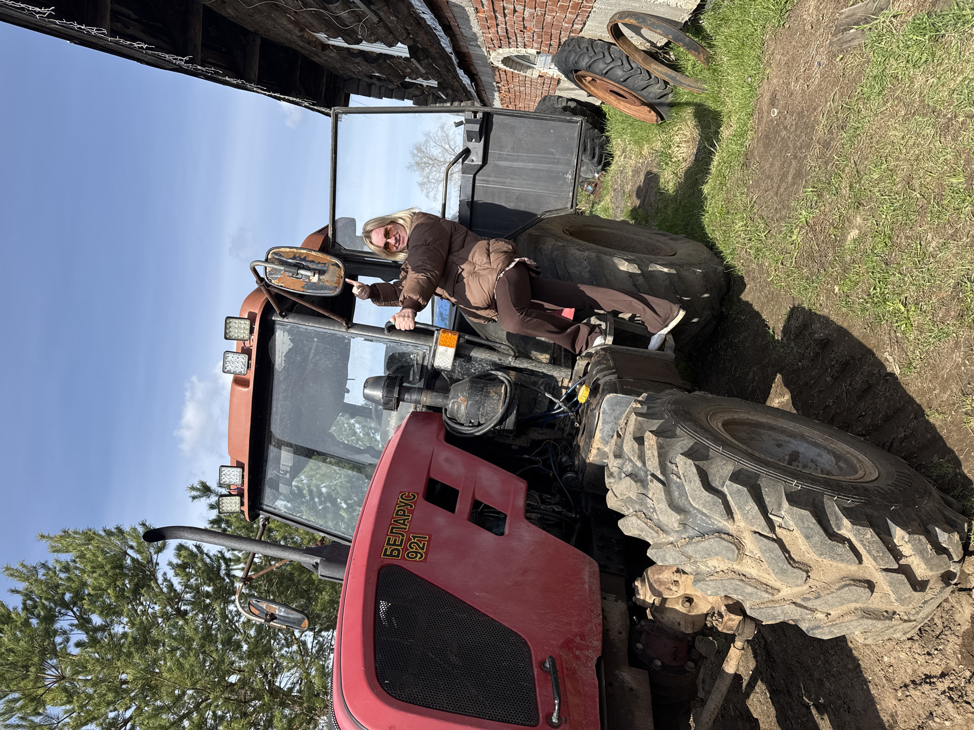 The image shows a woman standing on the tire of a red Belarus 921 tractor. The tractor is located outdoors, in a grassy area near a brick building and another wooden structure. Two spare tractor tires are sitting nearby. The woman is wearing a brown jacket and pants and white sneakers. She is pointing to the left. The tractor has lights on top and a mirror attached to the side. The location is Уральское, Russia.