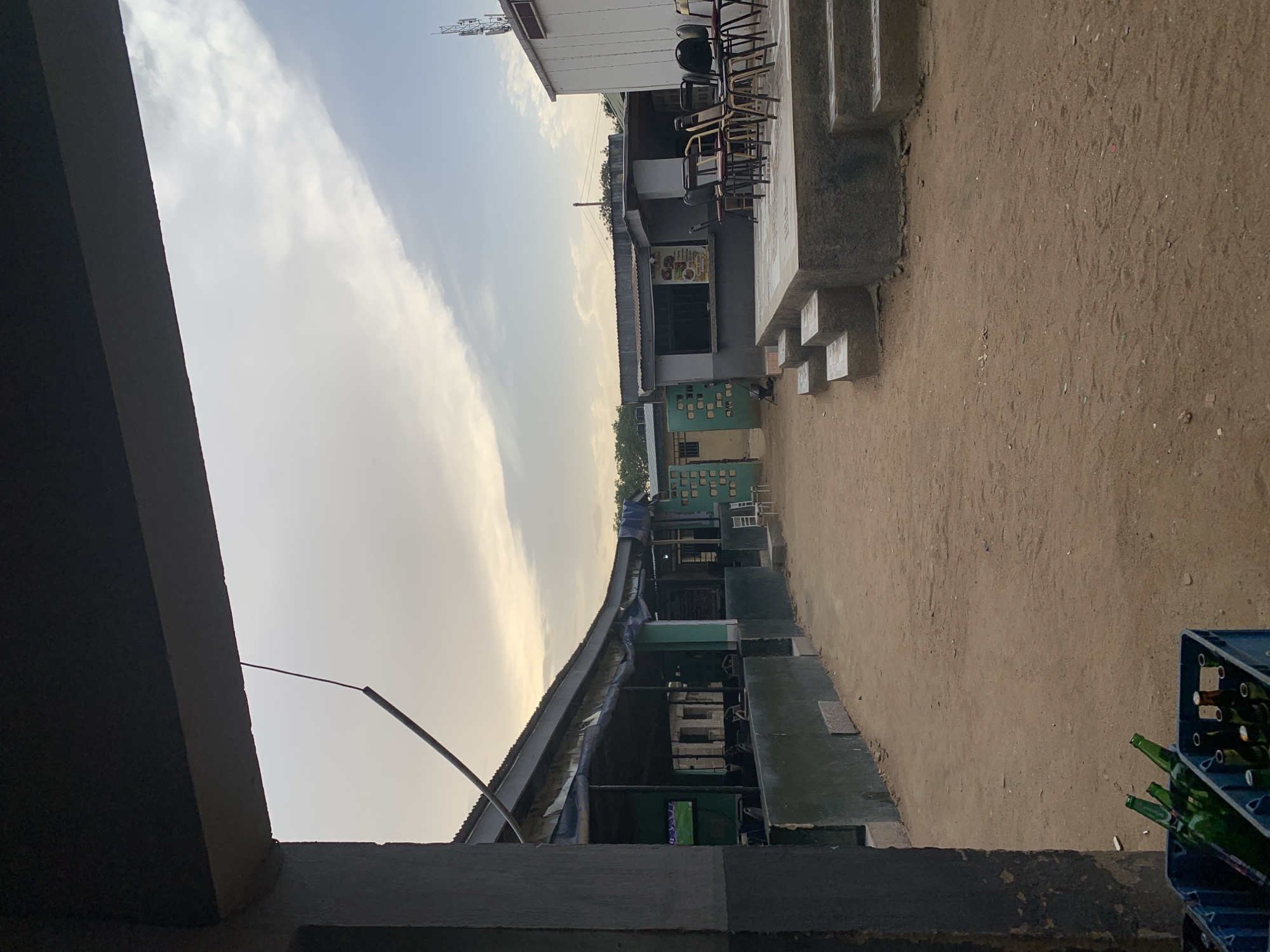 This outdoor shot shows a street in New Stadium, Nigeria. The ground is dirt and lined with buildings. The buildings are painted in greens and whites. One structure is an overhang. Crates of empty green glass bottles are in the foreground. The sky is light with some clouds.