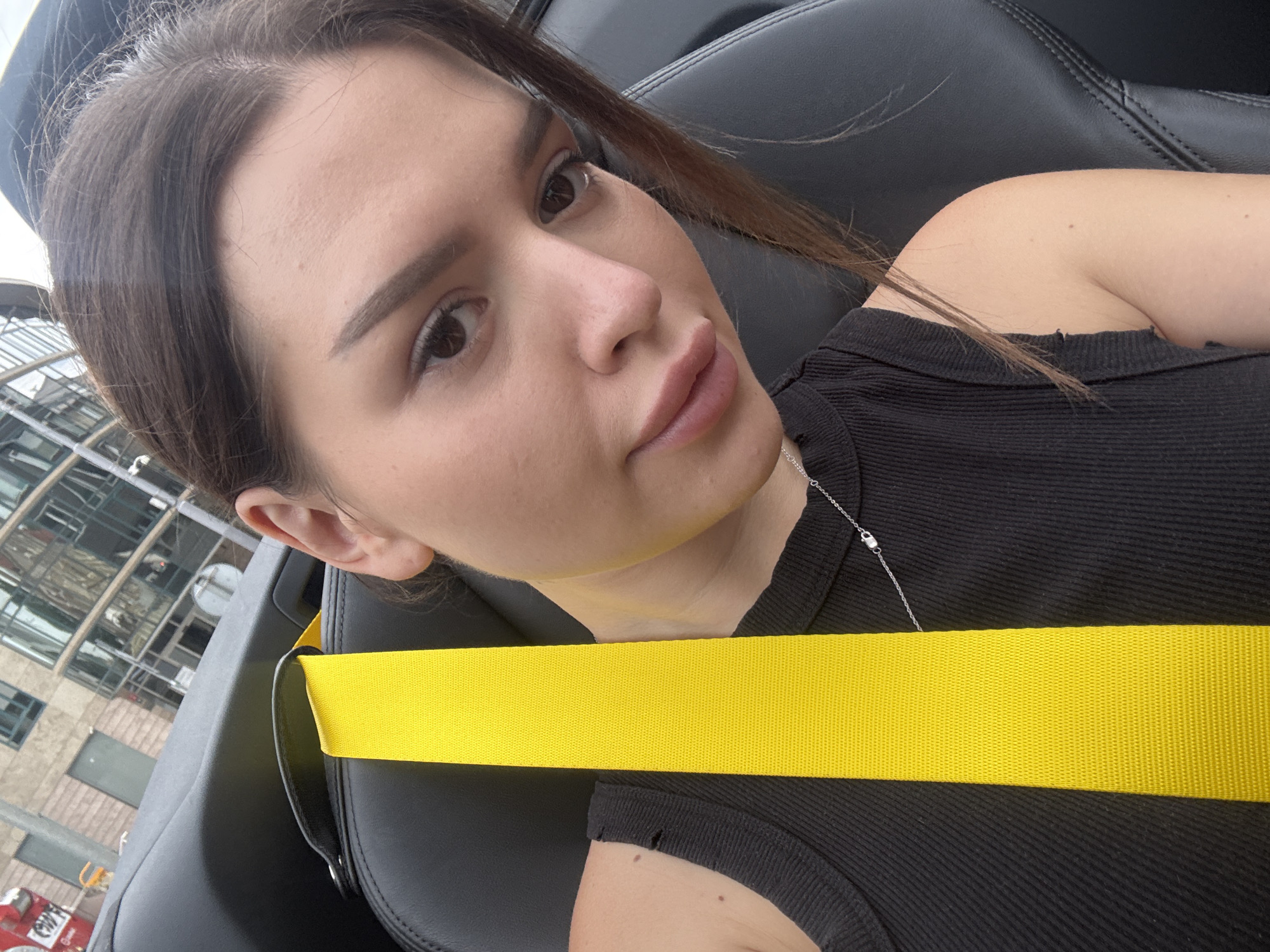 A medium shot is captured from a first-person perspective, looking down and slightly to the right at a young woman seated in a car. She has her right arm resting on the seat and is looking directly at the camera. The woman has brown hair, brown eyes, and is wearing a black ribbed tank top and a silver necklace. A bright yellow seatbelt runs vertically down the center of the frame, over her shoulder and chest. The background shows a blurred view of a modern building with large windows. The interior of the car, with black leather seats, is visible. The scene appears to be in a city environment, likely Prague, Czechia, based on the architectural style of the building in the background.