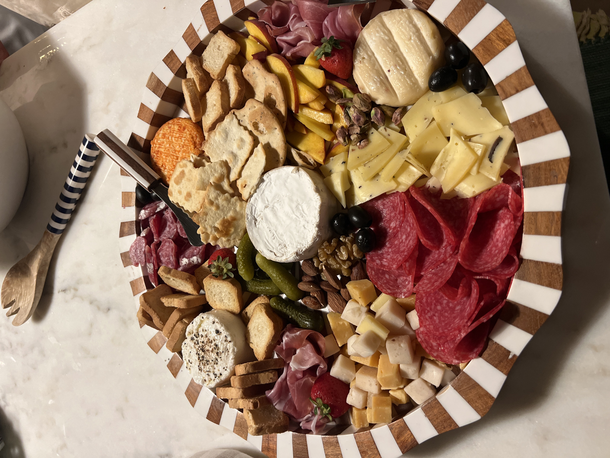 The image shows a charcuterie board arranged on a marble countertop. The board is round and has a distinctive striped rim. It is filled with various food items, including cheeses, cured meats, crackers, fruit, nuts, and pickles. Specifically visible are: a round cheese with black pepper, a wheel of brie, a bright orange cheese, sliced salami, prosciutto, diced cheeses, strawberries, sliced peaches, olives, almonds, pistachios, small pickles, and various types of crackers and breadsticks. A wooden salad fork with a striped handle rests on the marble surface near the board. The lighting suggests an indoor setting, possibly a kitchen or dining area. There are no people or animals visible in the image. The location in Vila Nova de Cerveira, Portugal is not discernible from the visual information provided in the image.
