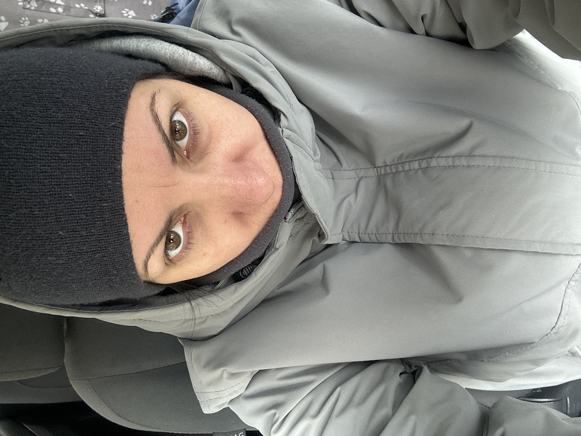 A woman with hazel eyes, wearing a black knit hat and gray hooded jacket, is looking directly at the camera. The setting appears to be inside a car, indicated by a black car seat visible in the lower left. There's no specific detail pointing to the location being Sofia, Bulgaria, so location data is not applicable.
