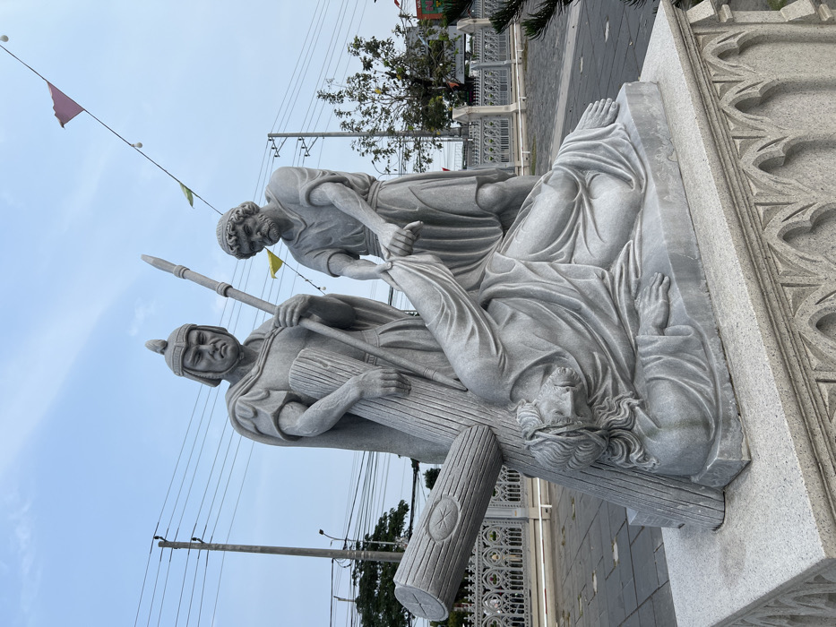 A large, detailed grey stone sculpture depicting three male figures from the Stations of the Cross is positioned outdoors on a dark grey paved area. The central figure, identified as Jesus, lies fallen on his left side, supported by his left hand, with a substantial wooden cross resting across his back and left shoulder. His face, with long hair and a beard, is turned upwards. To his immediate right, a bearded man in a simple tunic grasps Jesus's left arm, seemingly in an attempt to assist him. To the left and slightly behind Jesus, a Roman soldier in a helmet and full armor stands upright, holding a spear vertically with his left hand, his expression stern. The sculpture rests on a multi-tiered, lighter grey stone pedestal featuring gothic architectural relief patterns.
In the background, an ornate, light-colored gothic-style fence runs horizontally. Beyond the fence, utility poles and numerous black electrical wires traverse the scene against a clear, light blue sky. Scattered green foliage from trees is visible, along with small triangular flags (yellow and pink) and strung globe lights in the upper right quadrant. The location is the outdoor grounds of Thành Đường Đức Mẹ Hằng Cứu Giúp (Our Lady of Perpetual Help Church) at 38 Kỳ Đồng, Ward 9, District 3, Ho Chi Minh City, Vietnam.