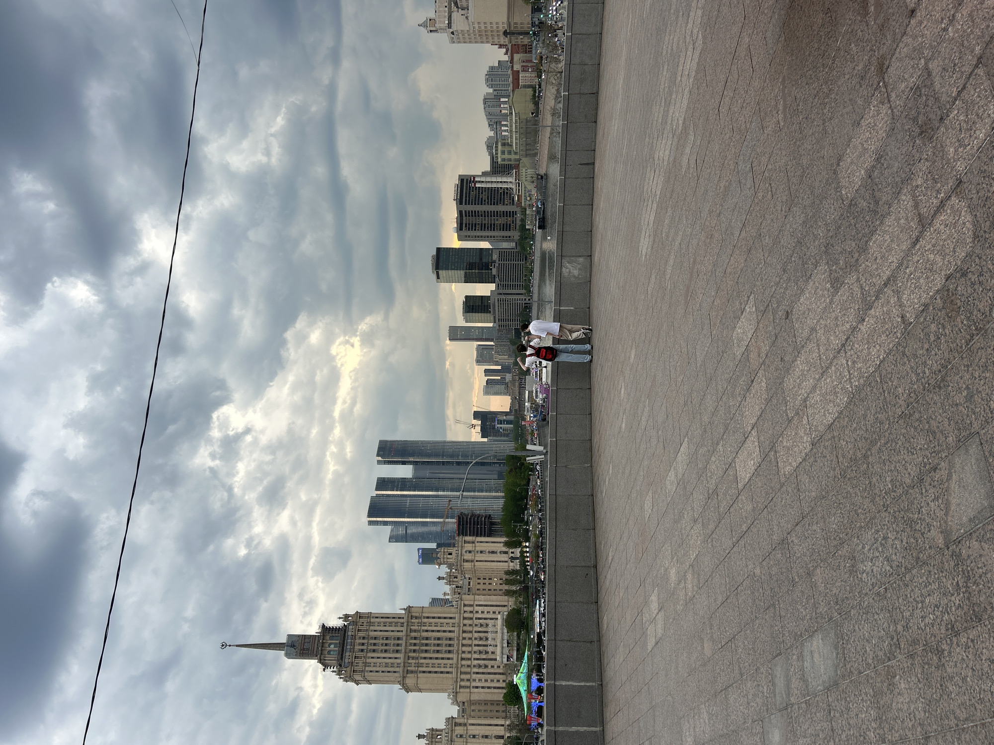 The image depicts a city scene under a cloudy sky. A tall building with a spire is visible in the lower left, indicating an older architectural style. Modern skyscrapers rise in the background. A paved walkway with stone blocks occupies the right side of the image. Two people stand on the walkway, their faces obscured. The environment includes visible street traffic and vegetation between the buildings. The location appears to be a city with a mix of old and modern architecture.