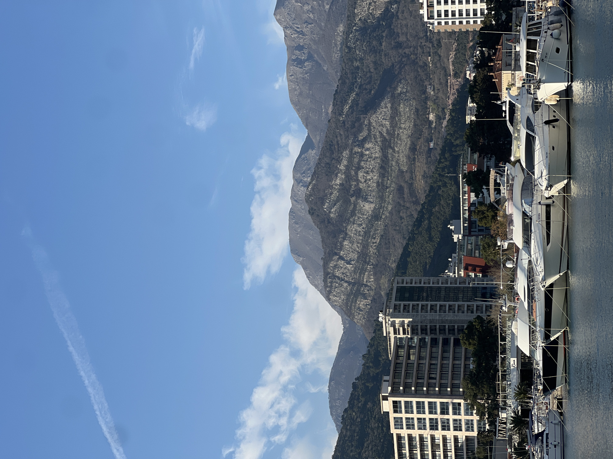 The image displays a marina in Budva, Montenegro. In the foreground, numerous luxury motor yachts and smaller recreational boats are moored on calm, dark blue water. These vessels are predominantly white with dark accents, featuring visible mooring lines and fenders. In the mid-ground, several modern multi-story buildings, appearing to be apartment complexes or hotels with numerous windows and balconies, line the waterfront. Green foliage from trees and bushes is interspersed among the structures, and a sign for "Kuzina" is partially visible. The background is dominated by a range of large, rugged mountains with varied rock formations and sparse vegetation, indicating a rocky, possibly deciduous terrain. The sky above is a clear, bright blue with scattered white clouds and a distinct white contrail stretching across the upper left quadrant. No individuals or specific human actions are discernible within the frame. The scene is illuminated by natural daylight, casting subtle shadows.