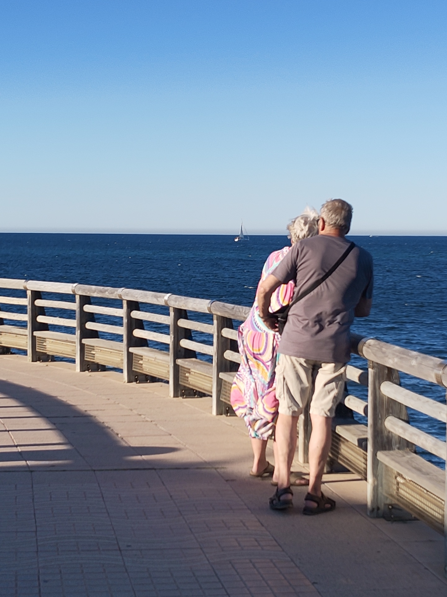 A photograph captures an elderly couple from behind as they stand on a pier, gazing out at the sea. The location is most likely the city of Dénia, Spain, given the pier's architecture, the Mediterranean Sea, and the clear blue sky. The pier is constructed with wooden railings and benches along its side, and the couple is leaning against it. The man is wearing a gray t-shirt, khaki shorts, and sandals, with a strap visible across his back. The woman, standing slightly behind him, wears a colorful, patterned dress and sandals. In the distance, a sailboat is visible on the sea. The tiled pier floor shows shadows from the railings and the couple. The sky is a clear, cloudless blue.