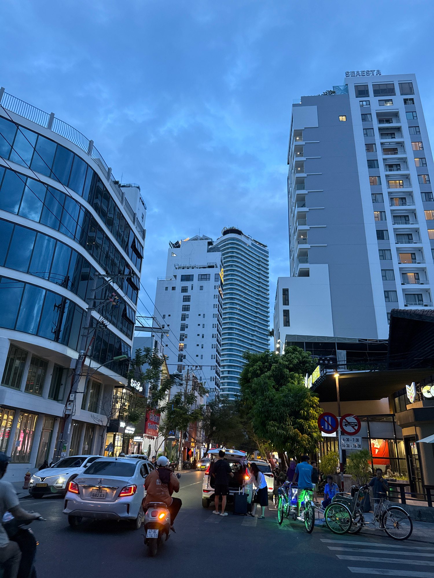 The image depicts a street scene in Nha Trang, Vietnam, likely during the evening or dusk, as indicated by the dim lighting and illuminated building windows. The scene is dominated by modern, multi-story buildings, including a prominent white and gray high-rise with the name "SEAEASTA" visible at the top. Another distinctive building features a curved, white facade with horizontal accents.
The street is bustling with activity. Several cars are present, including a white sedan with its headlights on and a silver sedan in the foreground with a license plate reading "91 G82.63". A person on a scooter is visible in the lower left corner, and another scooter with two passengers is in the center foreground. Several people are gathered on the sidewalk, some near parked vehicles and others near what appears to be a cafe or shop. Two pedicabs, one with green illuminated accents, are parked on the side of the street.
Street signs are visible, including a traffic sign indicating restrictions on parking and a sign with times in Vietnamese, likely related to a schedule. Trees and vegetation are scattered along the street, providing some greenery amidst the urban environment. The sky is overcast with clouds. The overall impression is a lively urban streetscape in a coastal city.