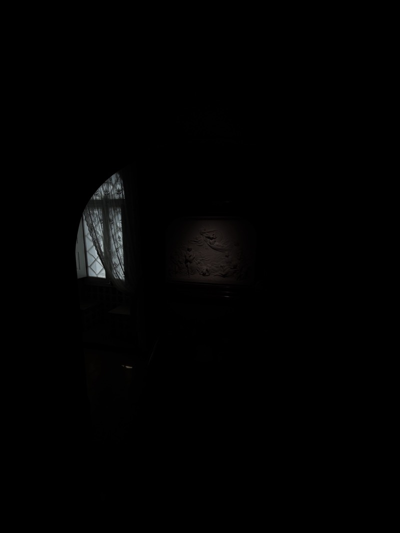 The image captures a dimly lit interior, possibly in Sintra, Portugal. A partially visible arched window with lace curtains is positioned to the left, allowing some light to enter the room. Adjacent to the window, a chair is silhouetted against the dark backdrop. Further to the right, a wall features a relief or mural, faintly illuminated. The overall environment is dark.