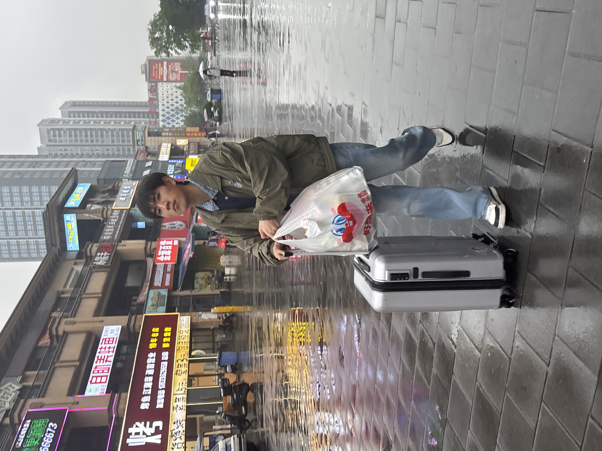 A young Asian male stands on a wet, grey brick sidewalk in Jiangjin, China, pulling a silver suitcase. He is wearing a green jacket, blue jeans, and dark shoes with white accents, and carrying a translucent plastic bag. The sidewalk reflects the surrounding environment, including neon signs and buildings. The backdrop features urban buildings and a wet street scene, suggesting it has been raining. Signs in Chinese are visible on the buildings, and a person with an umbrella is further down the street.