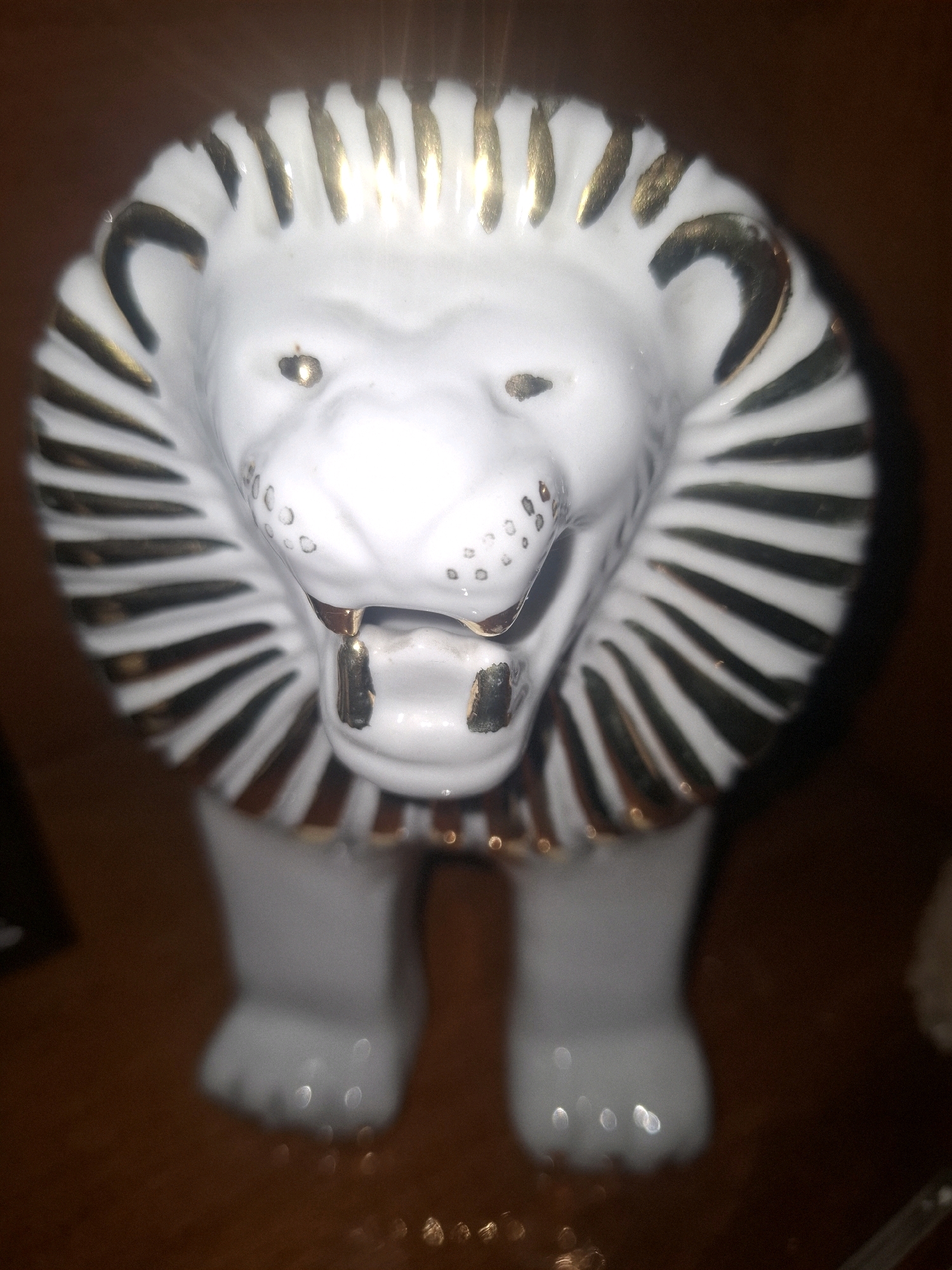 A ceramic lion figurine with a white body and a gold-striped mane stands on a brown wooden surface. The figurine has a detailed face with open mouth, gold-tipped fangs, and gold-accented eyes. The mane is composed of black and gold stripes radiating outwards from the head. The figurine's front paws are visible, and it is positioned centrally in the frame. The background is dark and blurred, with some out-of-focus reflections visible on the surface. The image does not contain any information about Almaty, Kazakhstan.