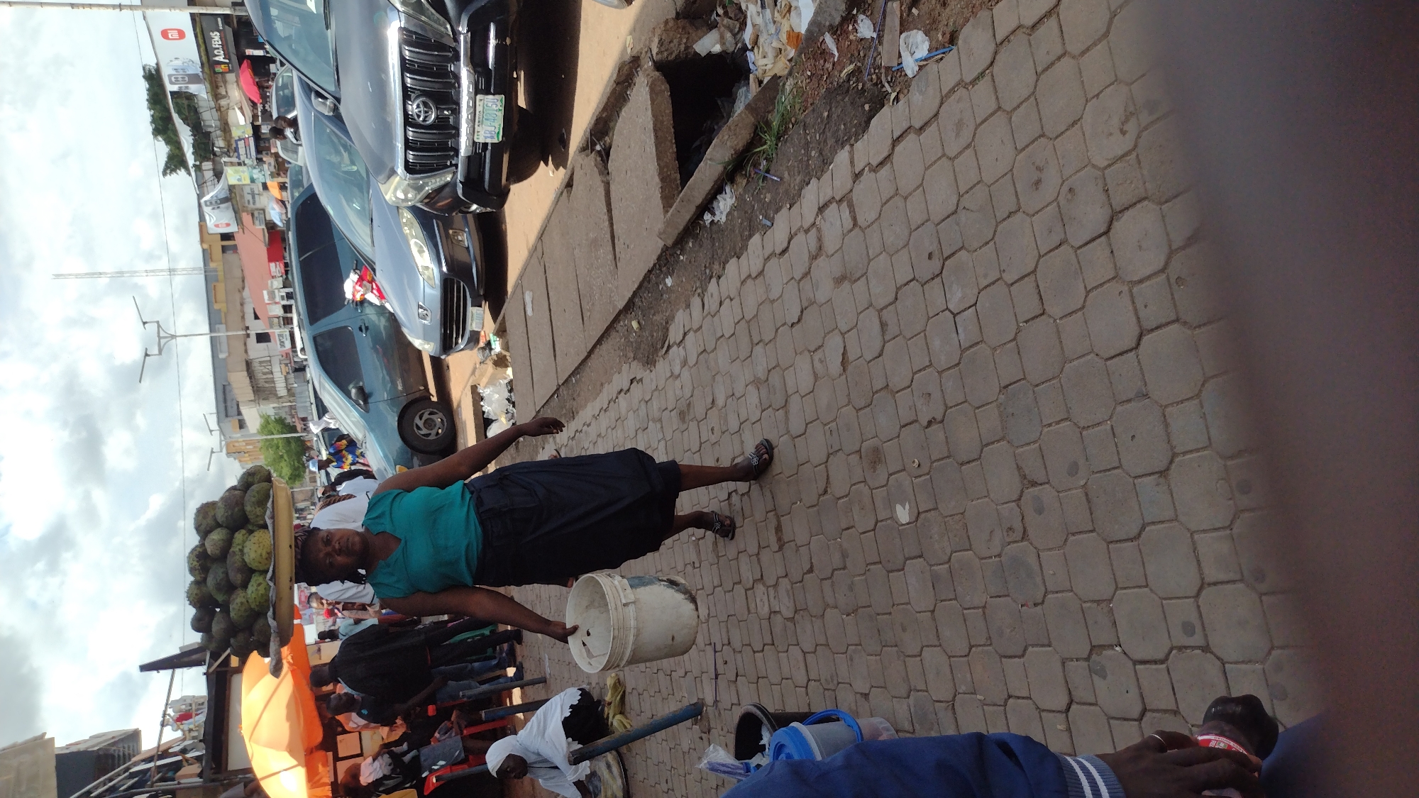 This is a photograph taken outdoors in Jos, Nigeria. The scene depicts a busy street market with a woman in the foreground carrying a large basket of fruit on her head. She is wearing a turquoise top and a dark skirt, and appears to be walking. To her left, a colorful umbrella provides shade. Behind her, several cars are parked, including a black SUV and a dark blue sedan. The ground is paved with interlocking bricks.
In the middle ground, other vendors and shoppers are visible, along with more parked vehicles and market stalls. There is a concrete drainage channel running alongside the paved area, with some debris and vegetation visible. In the background, buildings and structures suggest an urban environment. The sky is partly cloudy, indicating a potentially warm day.
The woman's expression is neutral, and the overall atmosphere seems to be one of daily commerce and activity. There are no explicit indications of a specific event, but rather a snapshot of a typical day in a Nigerian market. A white bucket is visible in the woman's hands. Some text is visible on a sign in the background which says "AO FEMS".
