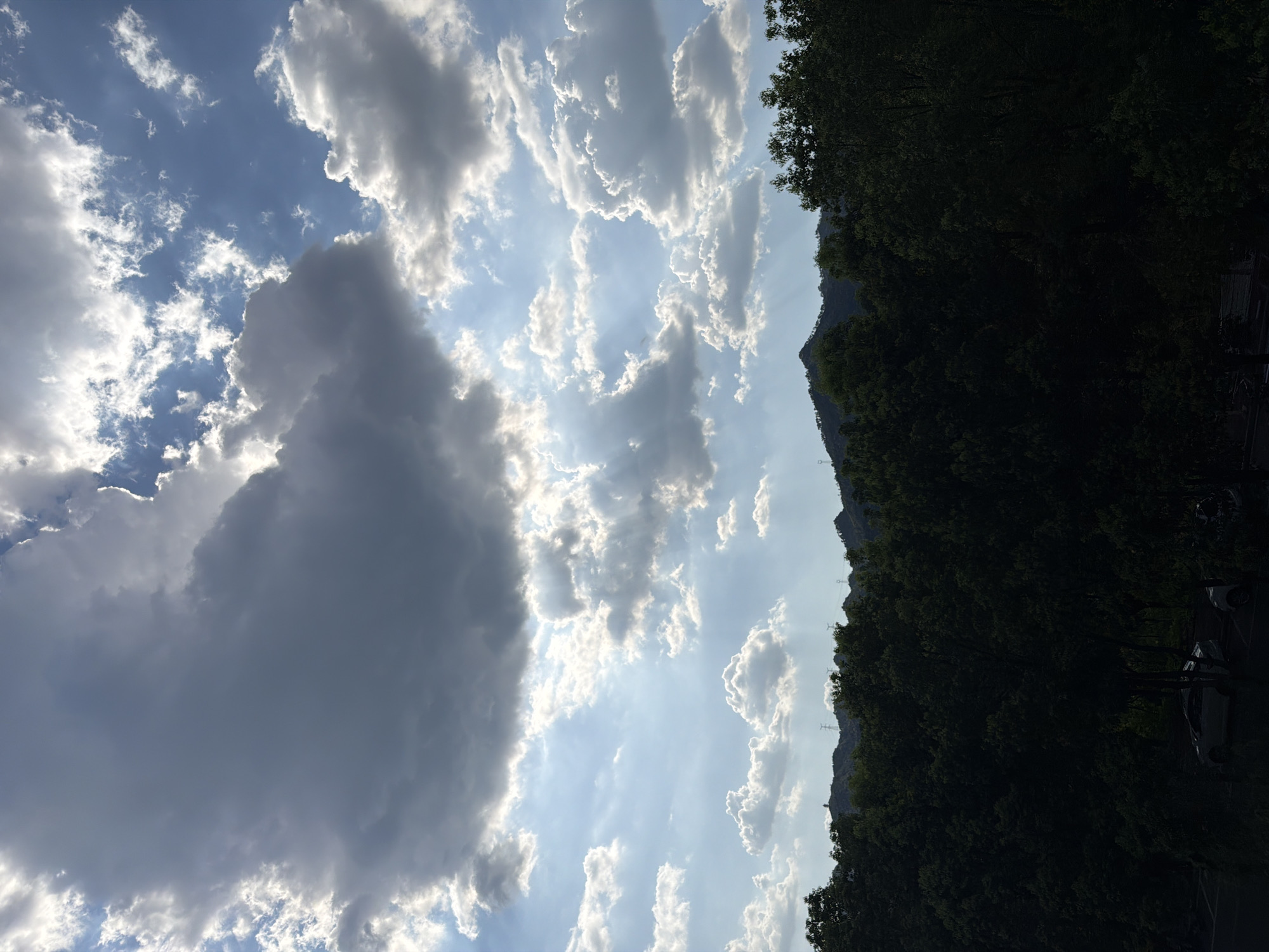This is a photograph of a sky with dramatic clouds and sunbeams filtering through them, above a dark silhouette of trees and mountains. The image is taken from a position looking upwards, with the lower portion of the frame dominated by the dense green foliage of trees, which are so dark they appear almost black. Beyond the trees, a range of hills or mountains is visible, with power lines and perhaps a distant structure on the peaks. The sky is a mix of bright blue and grey clouds, with shafts of sunlight piercing through the darker clouds, creating a dramatic, ethereal effect. Some of the clouds are puffy and white, while others are dark and brooding. The overall impression is one of natural beauty and atmospheric drama, likely captured at either sunrise or sunset. There are no people clearly visible in the photograph. The scene is not specifically identifiable as being in Jiangjin, China, but it depicts a natural landscape consistent with many locations worldwide.