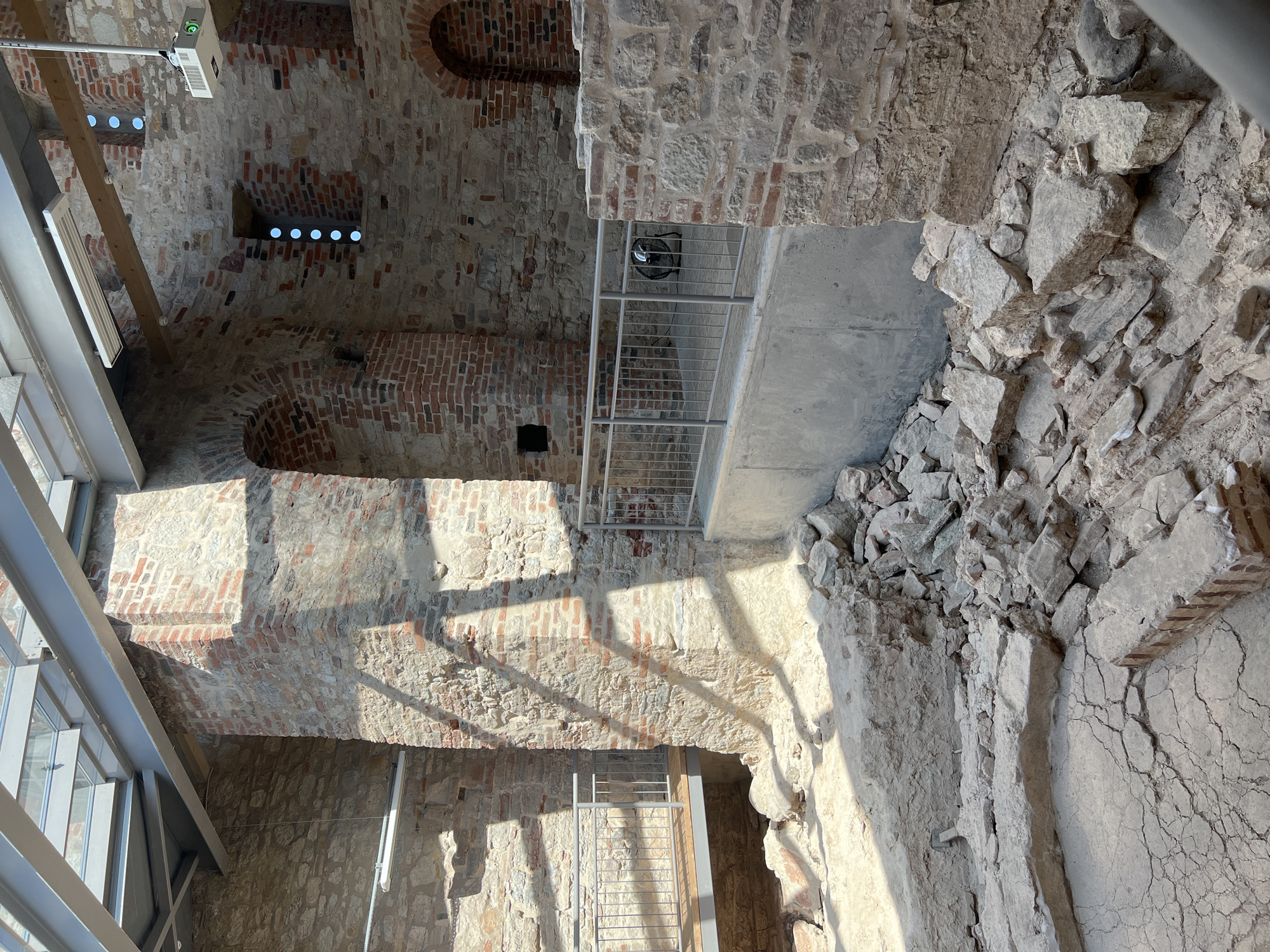 The image depicts the interior of an archaeological site, likely a historical building undergoing preservation or exhibition. The scene is characterized by exposed ancient brick and stone walls, some of which are partially reconstructed or stabilized with modern concrete elements. A glass roof structure with metal beams is visible overhead, suggesting the site is enclosed for protection. In the foreground, there are piles of rubble and cracked earth, indicating an excavation or ruin. Modern safety features such as metal railings and a walkway are integrated into the structure, allowing visitors to observe the archaeological remains. The lighting suggests daytime, with shadows cast by the roof structure. Based on the context of archaeological preservation and historical architecture, this scene is likely located within the city of Veliky Novgorod, Russia, a city renowned for its extensive historical and archaeological sites. No individuals are present in the image.