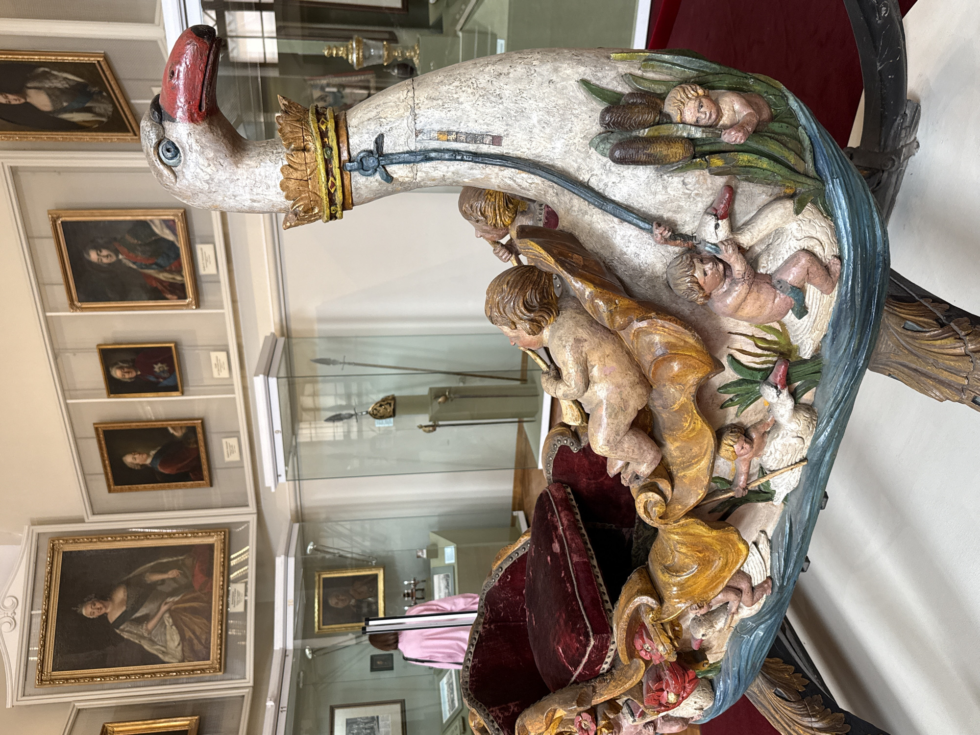 The image depicts a decorative wooden swan sculpture, likely a carriage or boat, with cherubic figures carved into its design. The swan has a white body, red beak, and a golden crown around its neck. The cherubs are carved in various poses, some interacting with the swan and its surroundings, which include reeds and water. A red velvet-cushioned seat is visible within the swan's body. The sculpture is displayed indoors, possibly in a museum, with other artifacts and paintings visible in the background. These include portraits in ornate frames and display cases containing historical items. A person wearing a pink jacket is seen in the background, looking at an exhibit. The setting suggests a cultural institution. While the exact location is not specified, the presence of the Russian flag in the background of the provided crop suggests it is in Moscow, Russia.
