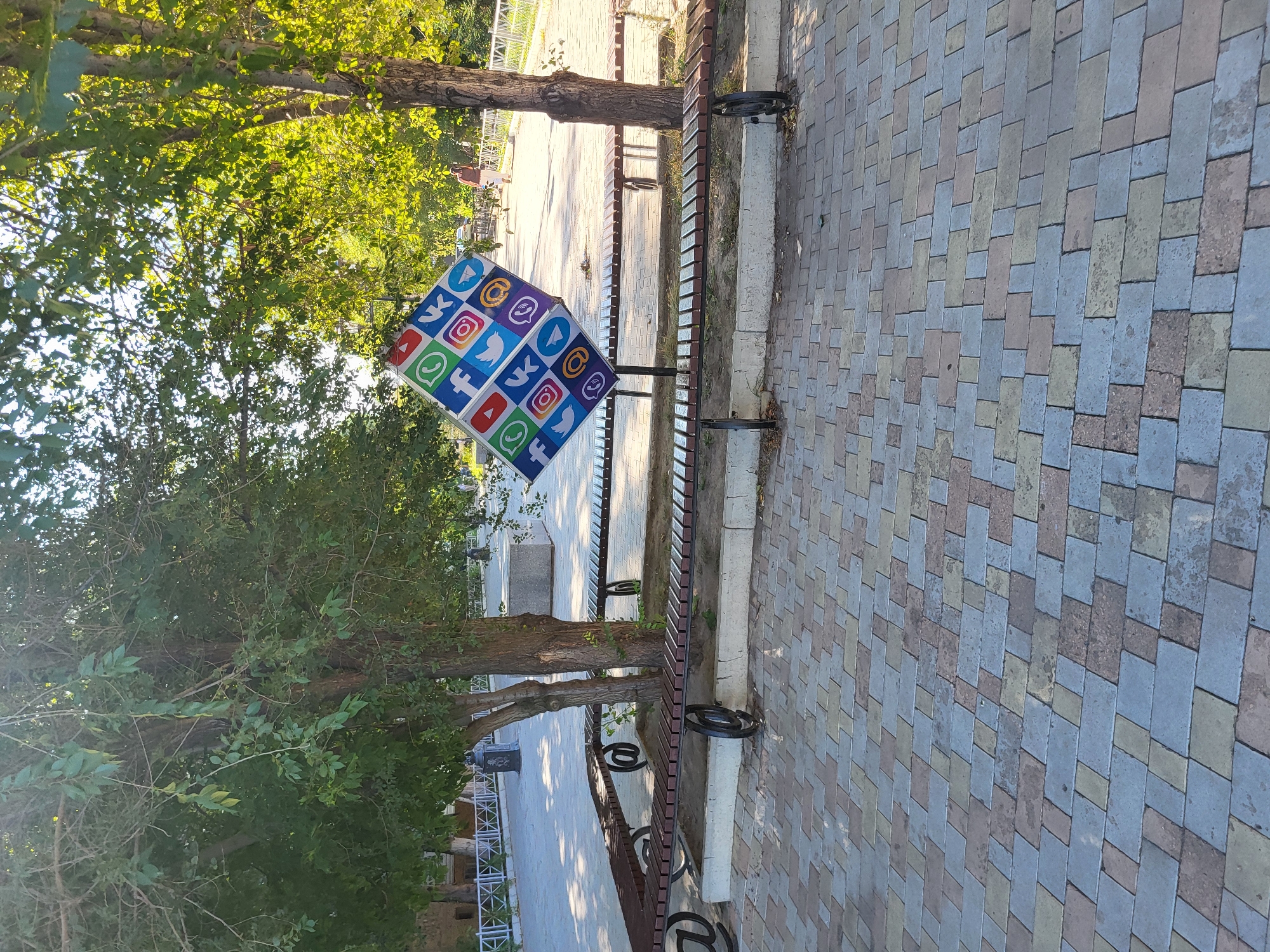 The image shows an outdoor public space in Petropavl, Kazakhstan, possibly a park or promenade. In the foreground, a paved walkway is made of multicolored rectangular bricks. Two long wooden benches with metal frames are visible, one in the foreground and another further back, both arranged around trees. A prominent feature is a large, diamond-shaped sign displaying logos of various social media platforms, tilted on a stand. Trees with green foliage are present, casting shadows on the ground. In the background, a paved area continues, with more trees and a white railing along the edge. A person is walking in the distance. The scene is illuminated by sunlight.