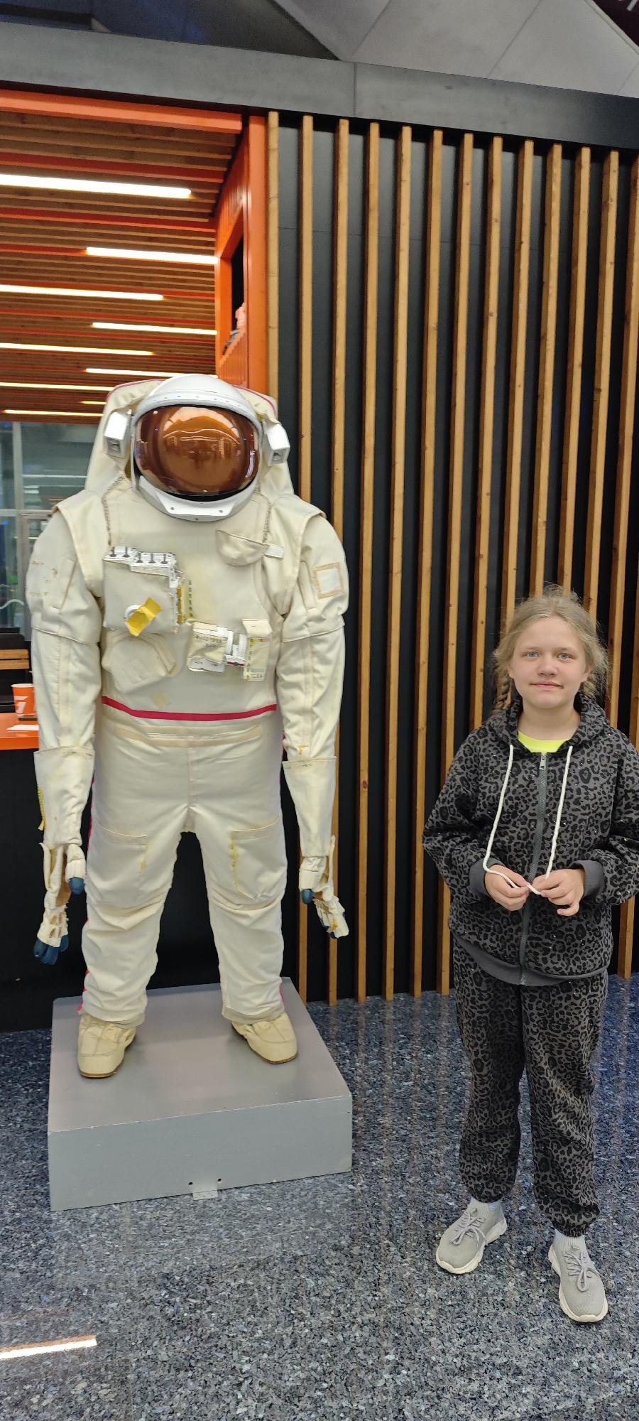A full shot captures a young girl standing next to a spacesuit display at the "Galaxy" cultural and entertainment center in the city of Estosadok, Russia. The spacesuit is white with a copper-colored helmet and various attachments and pockets. The girl is wearing a gray leopard-print hoodie and matching pants with gray sneakers. Behind them is a wall of vertical wood slats alternating in light and dark tones. The floor is a speckled gray surface.