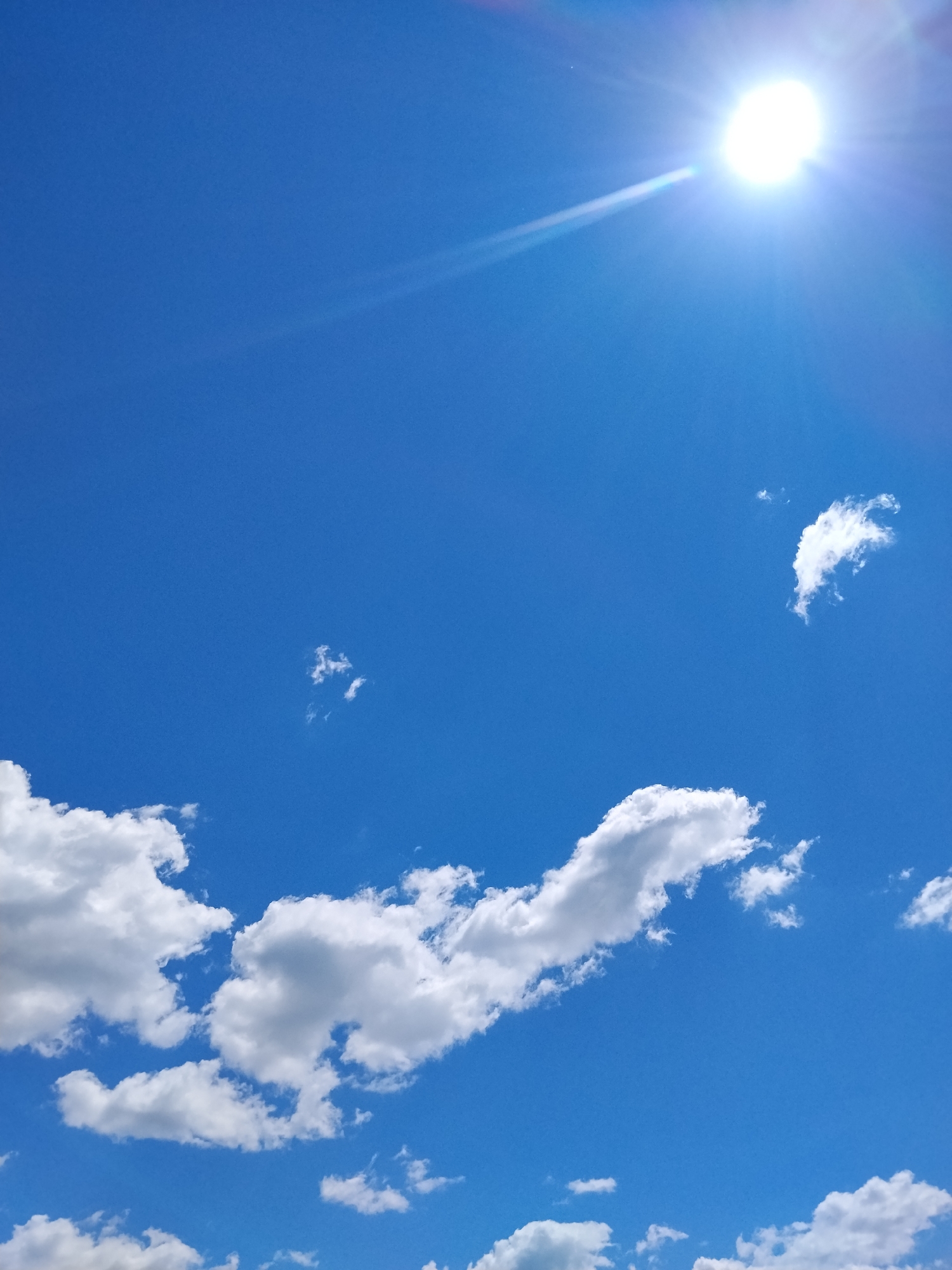 This is a vertical, eye-level, medium shot of a bright sunny sky with scattered clouds. The sun is high in the upper right corner, emitting a strong glare. The sky is a clear, vibrant blue. In the lower half of the image, there are several fluffy white cumulus clouds. The largest concentration of clouds is in the bottom left, with smaller, more dispersed clouds scattered throughout the frame. There are no people or man-made objects visible in this image, which is a natural scene. The scene is typical of a clear day in Ustyvytsia, Ukraine, likely during daylight hours.