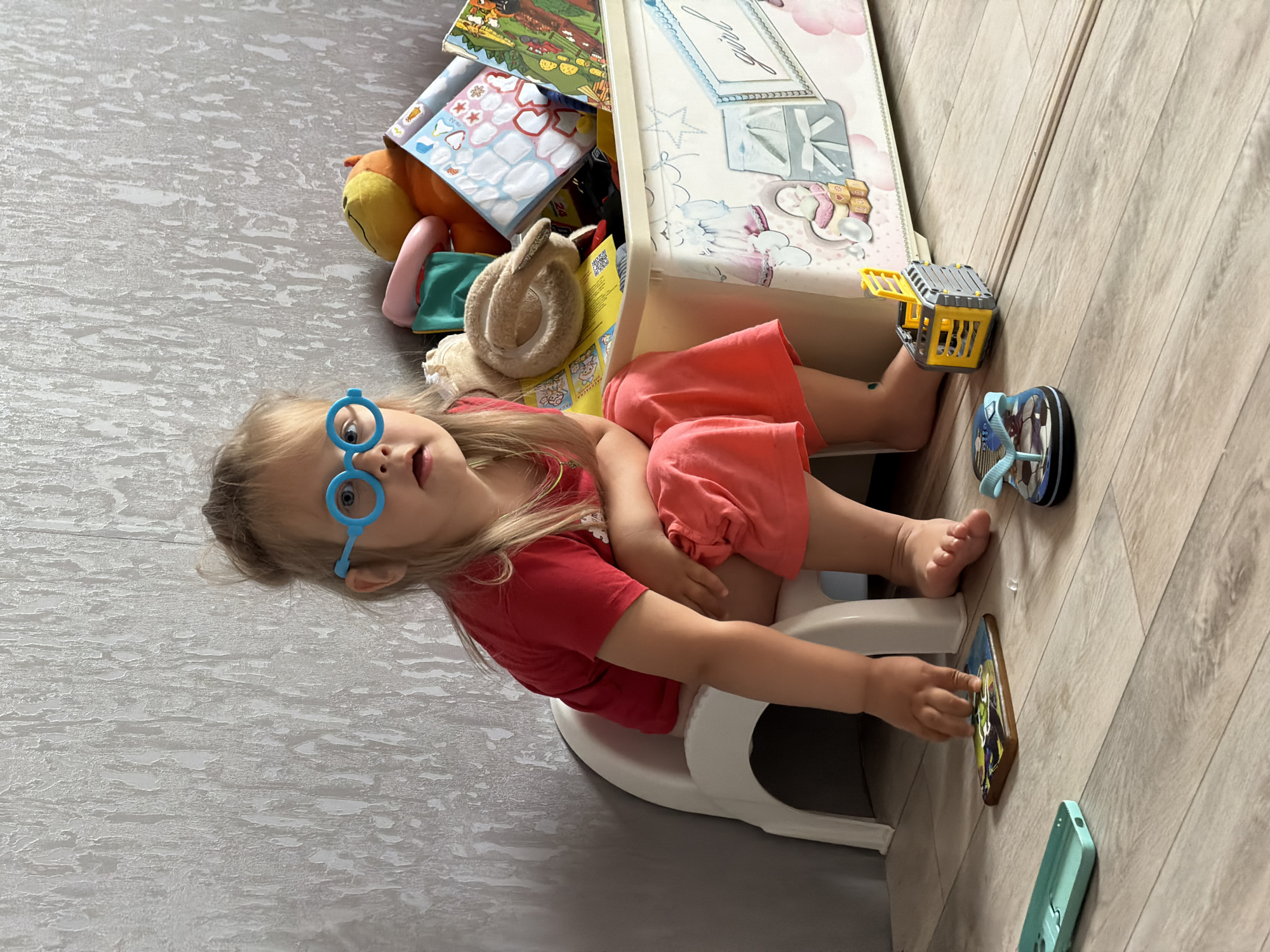 A young girl wearing blue circular glasses and a red t-shirt sits on a white potty chair. She is wearing coral-colored shorts. To her right, a toy storage bin is filled with stuffed animals and books. In front of her, on the floor, are a blue flip-flop, a small yellow toy construction vehicle, a phone case, and a small wooden object with an image on it. The scene appears to be indoors, with textured wallpaper on the wall behind the girl and a wooden floor. The location is not discernible from the image.