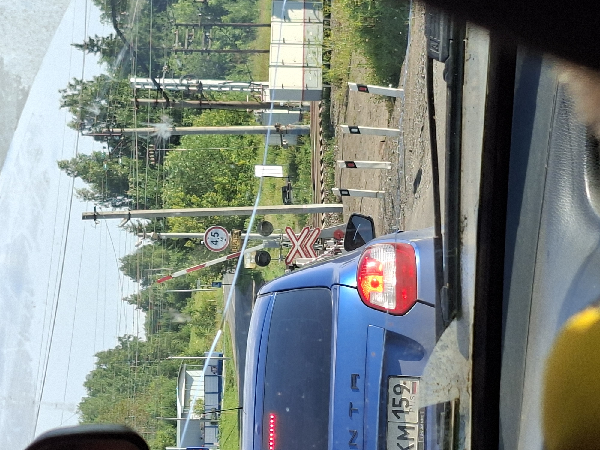 A blue sedan is stopped at a railroad crossing in Vereshchagino, Russia. The car's brake lights are illuminated. The railroad crossing has a barrier arm that is partially raised, and a red and white striped signal. A circular sign with a "4.5" and "M" in it is visible on a pole. The background consists of trees, power lines, and a building. The image appears to be taken from inside another vehicle.
