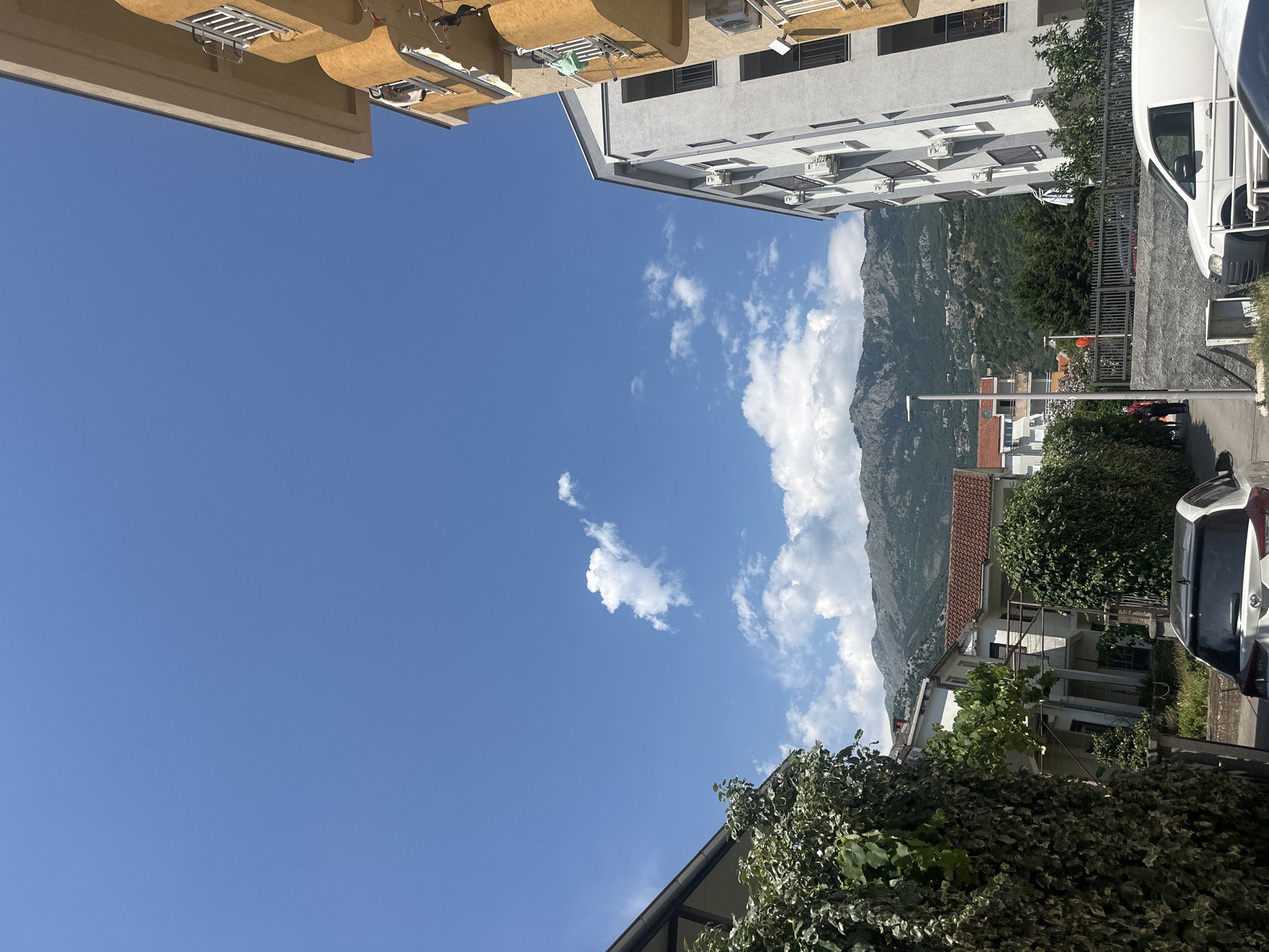 The image captures a view of residential buildings and a mountainous backdrop under a clear blue sky with scattered white clouds. In the foreground, on the left, a lush green ivy-covered wall dominates the frame, with the corner of a building with a dark tiled roof visible behind it. To the right, a modern white apartment building with multiple air conditioning units on its facade stands tall, with balconies featuring dark railings. Partially visible on the far right is another building with an orange-yellow facade, also with balconies.
In the mid-ground, a street runs between the buildings, with a white van and part of a white car parked along it. Several people are walking on the street. Behind the street, a low grey stone wall with a metal fence separates it from a grassy area with trees. Further back, several other residential buildings with red tiled roofs are visible. The background is dominated by a large, imposing mountain range with a rugged, uneven surface, sparsely covered with green vegetation. The overall impression is of a sunny day in a Mediterranean coastal town, possibly Budva, Montenegro, given the architectural style and the mountainous terrain meeting the sea.