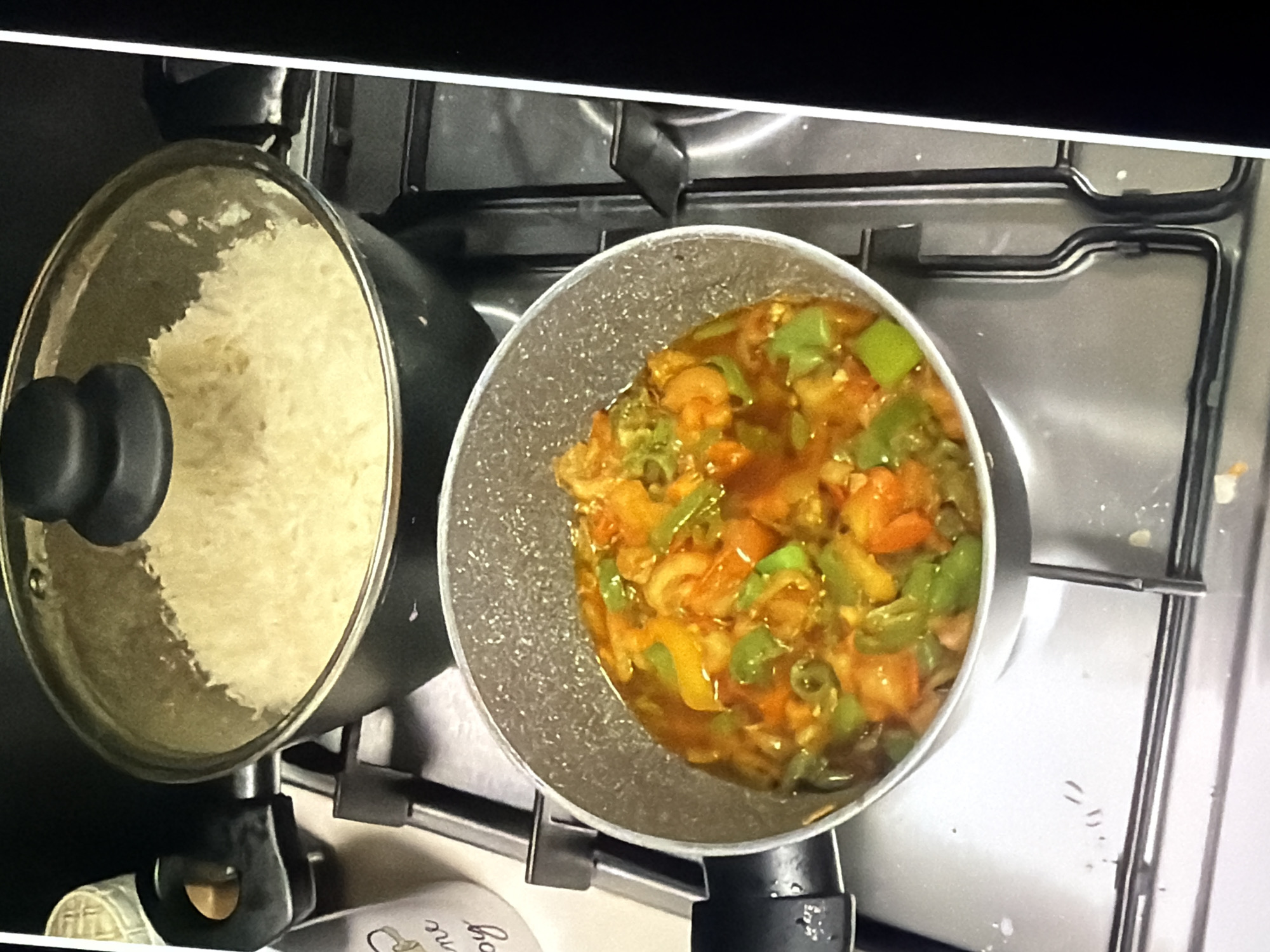 An overhead shot shows two pots on a stovetop. The pot on the left, in the foreground, is a small, grey pot filled with a vibrant mixture of orange and green vegetables in a reddish sauce. The pot on the right, in the background, is larger and black, with a clear glass lid. Inside this pot, a generous amount of fluffy white rice is visible. The stovetop is metallic and appears to be part of a domestic kitchen. To the left of the pots, a white cylindrical object with text on it is partially visible.
