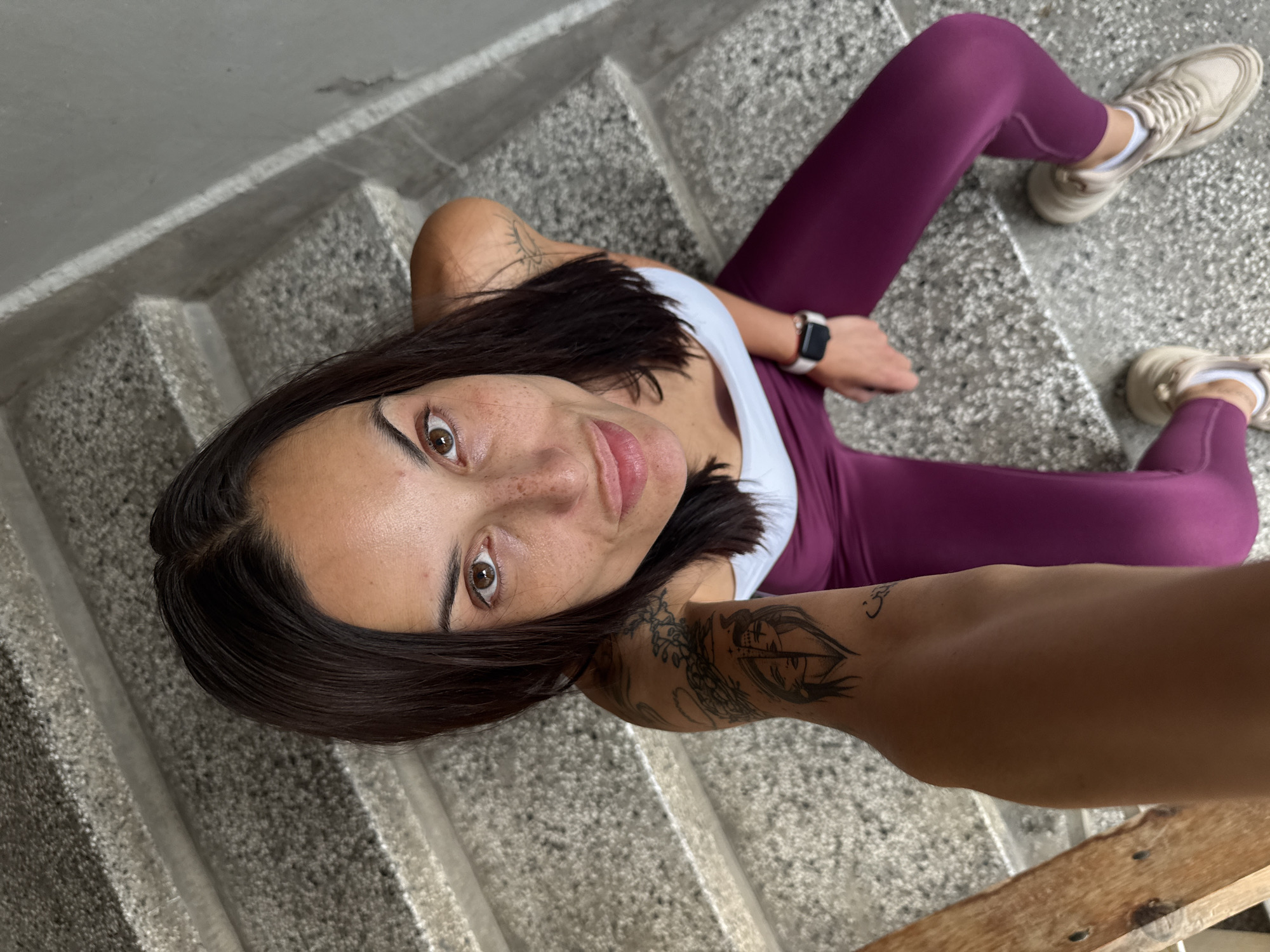 A woman is captured in a high-angle selfie, seated on a flight of light grey concrete stairs in Pleven, Bulgaria. She has dark hair, brown eyes, and visible freckles on her face, looking directly into the camera with a neutral to slightly inquisitive expression.
She is wearing a white tank top and dark purple (maroon) leggings, paired with light-colored sneakers and white socks. A smartwatch with a white band is on her left wrist. Tattoos are visible on both arms: a geometric or abstract design on her left shoulder/upper arm, and a more detailed artistic design, possibly featuring an eye motif and other elements, on her right forearm. A small smiley-like symbol is also visible higher on her right arm.
The stairs have a coarse, speckled texture. A wooden plank or similar object is partially visible in the bottom right of the frame. The lighting is soft and diffused, suggesting an indoor or covered setting, making the time of day undeterminable. No explicit text is clearly legible in the image, beyond the unidentifiable digital display on her smartwatch.