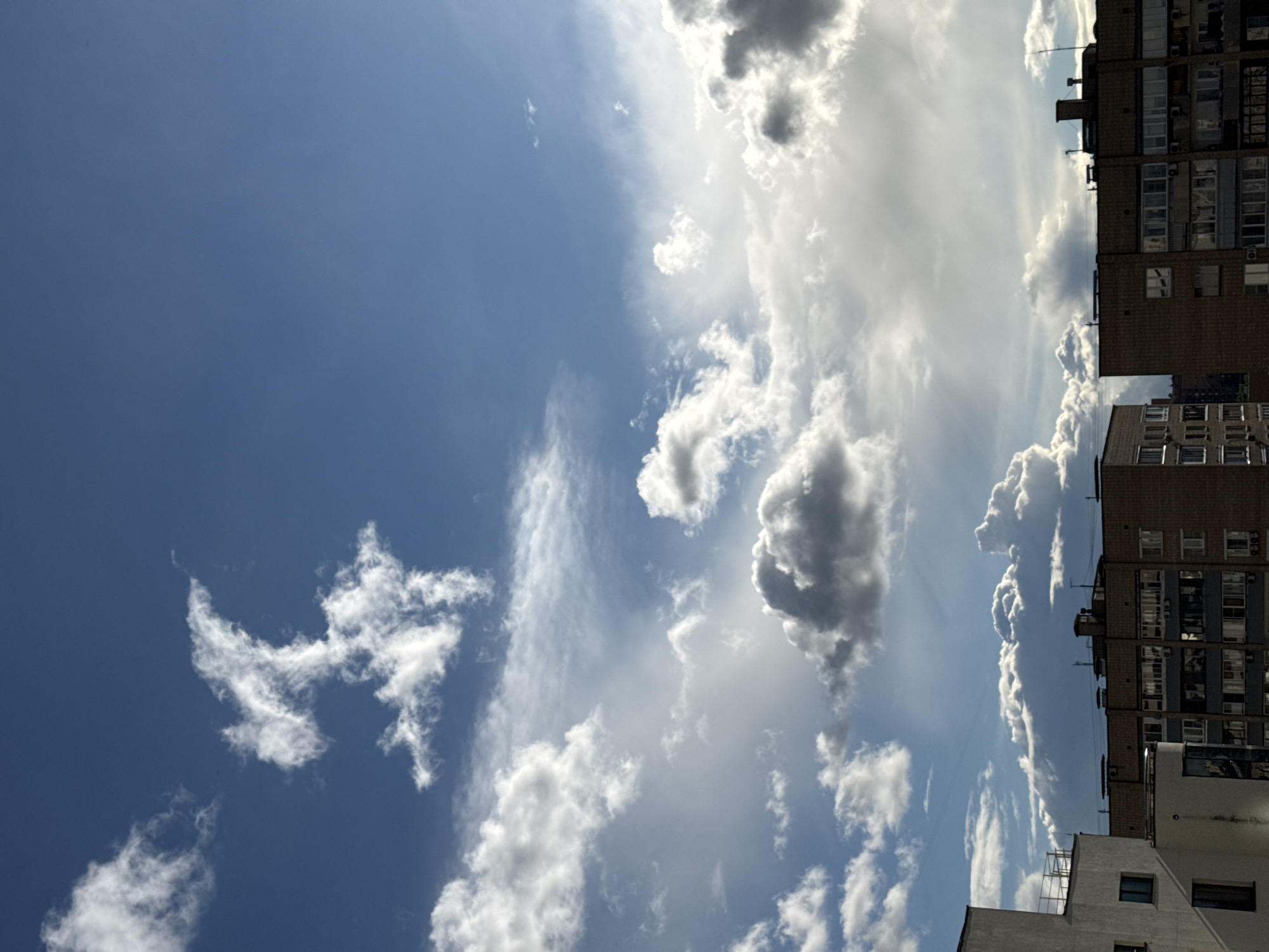The image captures a sky with scattered clouds of varying densities, ranging from thin wisps to dense, dark masses. A blue expanse is visible between the clouds. On the right side of the frame, portions of buildings are visible, showing architectural details such as windows and varying levels. The buildings are typical of urban residential structures, possibly indicating a location in a city. Given the prompt’s specification, the city is likely Moscow, Russia, and the buildings represent typical Moscow architecture, especially Soviet-era apartment blocks. No people or specific activities are directly visible in the frame.