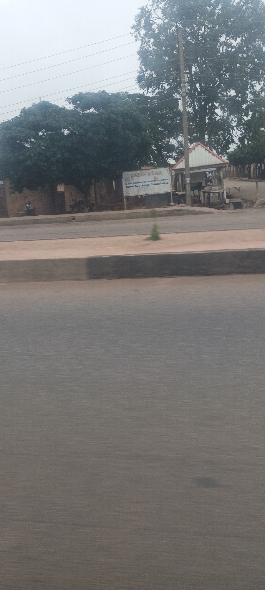 This media file captures a roadside scene, likely from a moving vehicle, indicated by the motion blur on the road surface in the foreground. The image is dominated by the multi-lane road and a concrete median barrier.
In the midground, beyond the road and curb, there's a dirt strip. Further back, a vibrant green tree with dense foliage occupies the left side. Beneath this tree, a light-brown building is visible, and a solitary figure is seated on a low structure or bench next to it, facing away from the camera.
Towards the center-right, a prominent white sign with blue lettering stands upright. The text on the sign reads:
"DADIN KOWA"
"To live together, we must live in peace"
"Zaman Tare Sai da Zaman Lafiya" (which is Hausa for "Living together requires peace").
To the right of the sign, a small, open-sided structure with a distinctive white roof and red trim, possibly a kiosk or a small shelter, is situated. A tall, large tree rises behind this structure, with multiple power lines traversing the overcast, grey sky.
The overall impression is of a quiet, possibly semi-urban or rural area during an overcast day, given the diffused light and lack of harsh shadows. The presence of Hausa text on the sign firmly places the location within a northern Nigerian context, consistent with Jos.