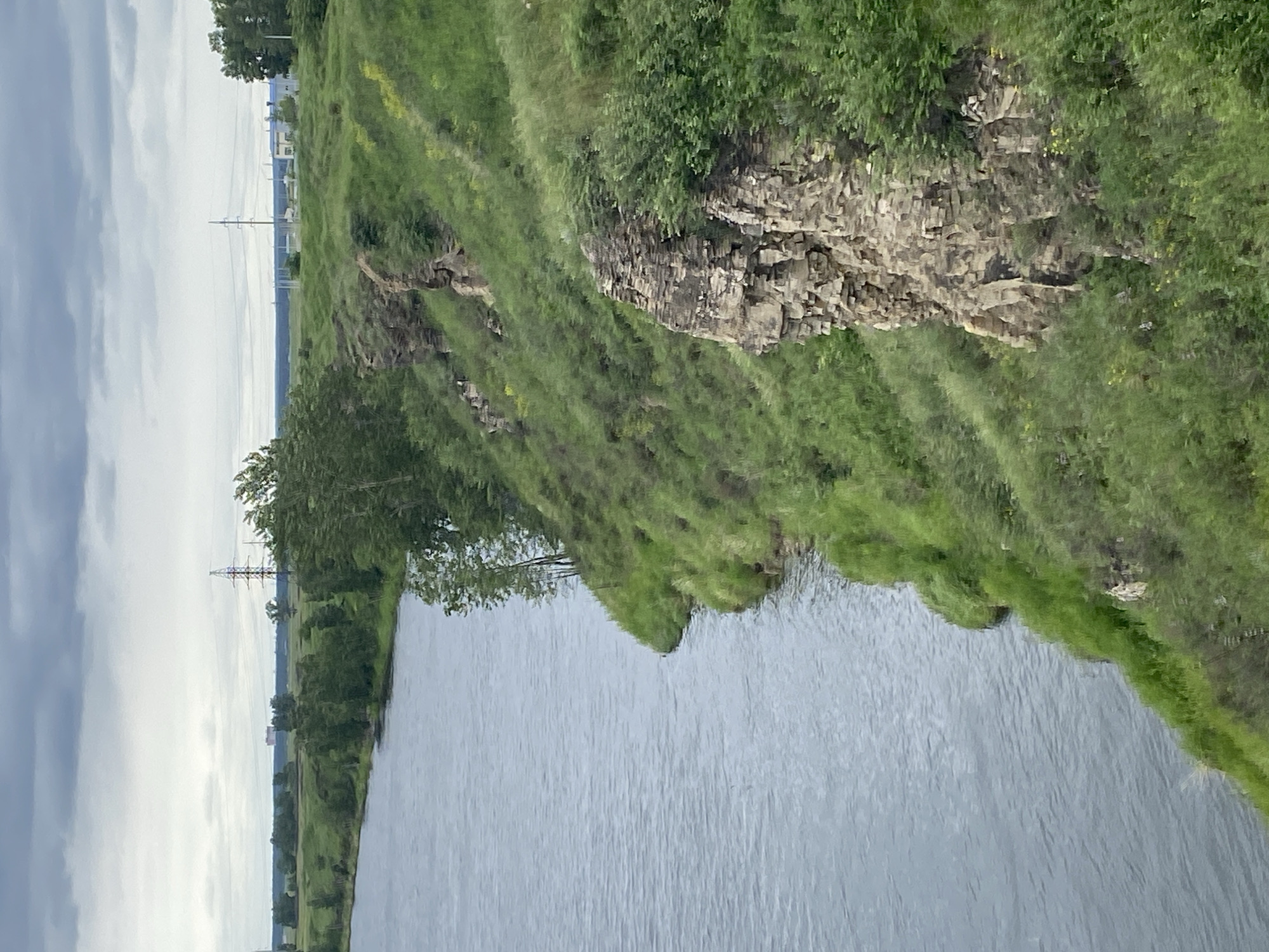 The image depicts a wide, calm river with a steep, grassy embankment on the right. The embankment features rocky outcrops and is covered in lush green vegetation, with scattered trees along the top and sides. In the distance, beyond the embankment and across the river, a flat landscape is visible with more trees and what appear to be industrial structures and power lines under a cloudy sky. The scene is likely located in or near Novomaltinsk, Russia, given the landscape and industrial elements often found in such regions. There are no people or animals visible in the image. The overall impression is a natural landscape with some human industrial presence in the background.