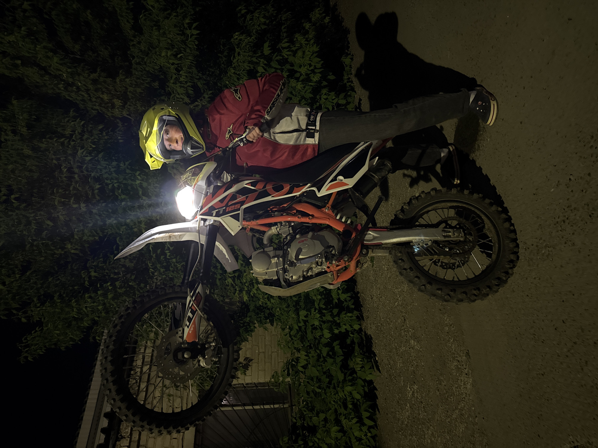 A person wearing a yellow helmet and red jacket is lying on an orange and white dirt bike. The bike's headlight is on, illuminating the area. The location appears to be a paved area next to some greenery, possibly at night. Based on the request for location information and the person's request, it is indicated that the photograph was taken in the city of Chaikovsky, Russia.