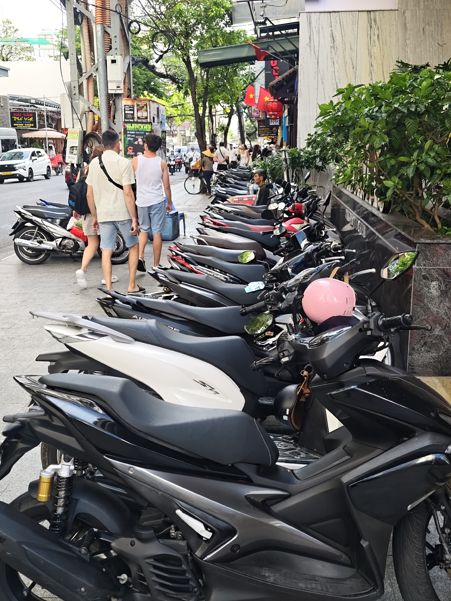 The image depicts a street scene in Nha Trang Ward, Vietnam, characterized by a row of parked motorbikes on a sidewalk adjacent to a building. Two men, one wearing a beige short-sleeved shirt and the other a white tank top, walk away from the camera along the sidewalk. They are followed by a person carrying a black backpack. In the foreground, several motorbikes are parked in neat rows, with a prominent pink helmet resting on the handlebars of one of the bikes. The background reveals more motorbikes, a white car on the street, and elements of urban infrastructure including utility poles and trees. The architecture suggests a commercial or residential area typical of a Vietnamese city.
