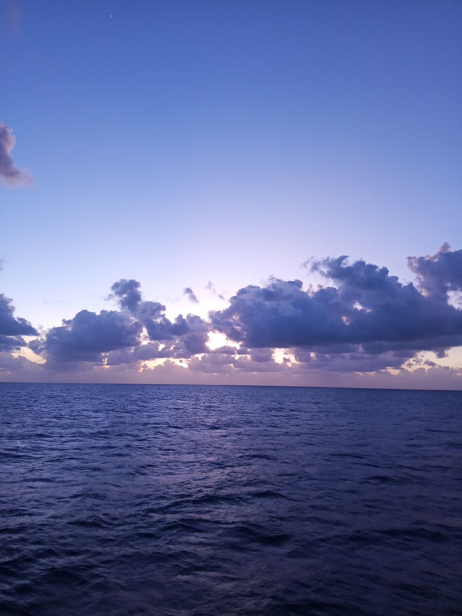 A twilight sky over the ocean features a crescent moon visible in the upper left quadrant. The sky transitions from a deep blue at the top to lighter shades of lavender and pink near the horizon, where scattered dark clouds are illuminated by the setting sun. The ocean, a deep blue, is marked by gentle waves and reflections of the sky. The scene is devoid of any human presence or man-made structures, suggesting a remote or natural setting. The overall impression is one of a serene and expansive seascape at dusk.