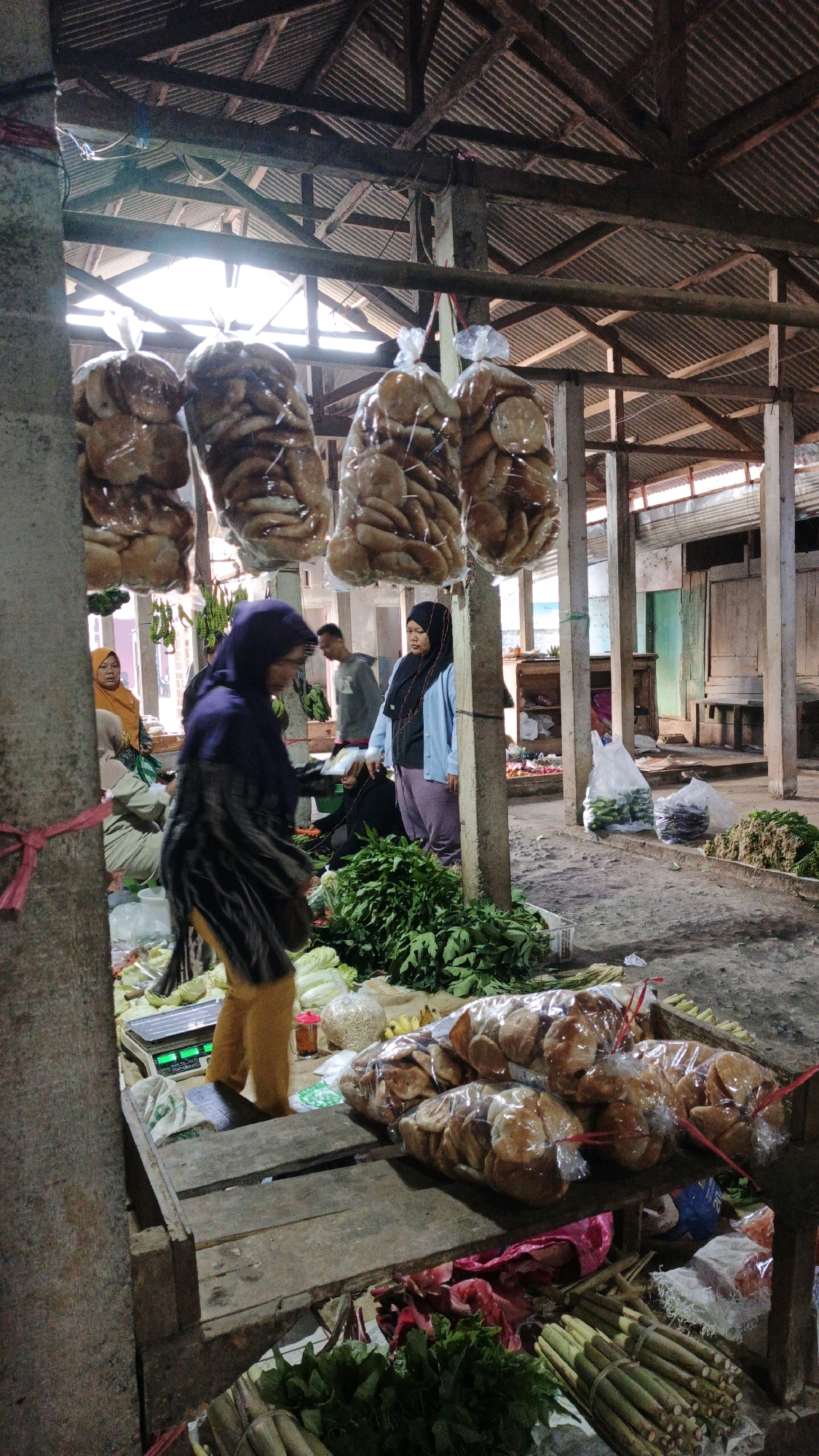 A bustling street market in Cibenda, Indonesia, is depicted. The scene is crowded with vendors and shoppers, with stalls overflowing with fresh produce and packaged goods. In the foreground, a woman in a purple headscarf and yellow pants is seen from behind, seemingly examining produce at a stall. Behind her, a pile of leafy green vegetables is visible, along with a digital scale.
Further back, several women in headscarves are engaged in conversations or tending to their stalls. A man in a grey hoodie stands between two women, one of whom is wearing a blue cardigan and light purple pants. Bags of biscuits hang from the ceiling in the middle ground. The market has a rustic feel, with a corrugated iron roof supported by wooden beams and columns. The ground is dirt and concrete. The atmosphere appears lively and communal, indicative of a typical Indonesian market.