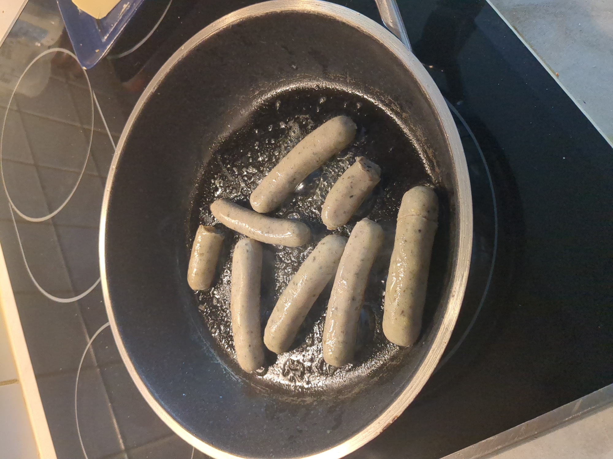 Eight sausages are being fried in a black, non-stick frying pan on a black ceramic stovetop. The sausages are light-colored and appear to be pre-cooked or partially cooked. The pan is filled with a small amount of oil, which is bubbling around the sausages. The stovetop has two circular heating elements visible. In the upper right corner of the image, a blue rectangular dish containing a slice of yellow butter is partially visible. The scene appears to be a kitchen. There is no information in the image that can be used to determine the location in the city of Nordhausen, Germany.