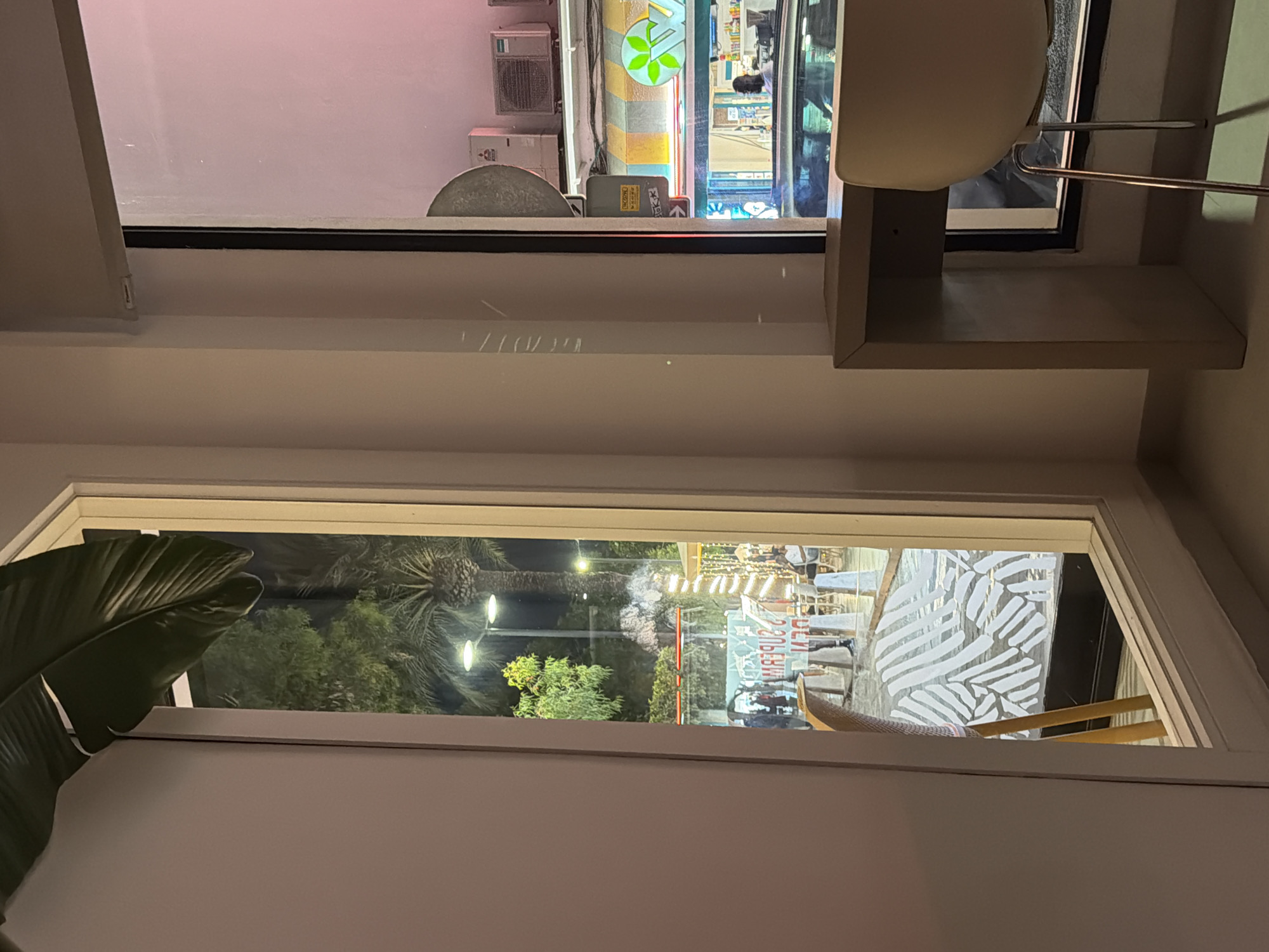 The image shows a view through a window at night. Inside, a white chair is visible. Outside, there is a street scene with palm trees and lights. Several people are walking in the distance. The reflection on the window shows the interior of a room with neon signs from a shop. One sign appears to be in English and the other in a non-Latin script. The overall impression is that of a bustling night scene in a tourist area.
