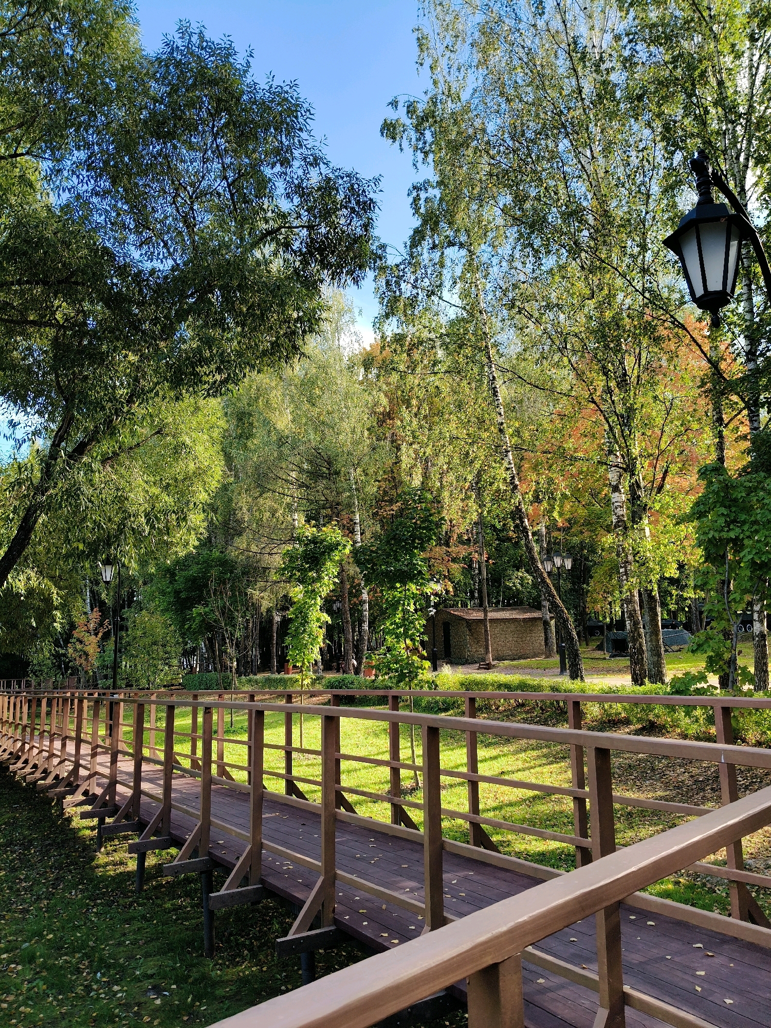 The image depicts a wooden bridge with a brown railing, set in a park-like environment. The bridge is elevated above a grassy area covered with fallen leaves. Trees with green and some autumnal foliage are visible in the background, along with a lamppost. There is a small brick building in the distance. The sky is blue and clear. The scene suggests an outdoor recreational area in Naro-Fominsk, Russia, possibly during the autumn season. No people are present in the image.