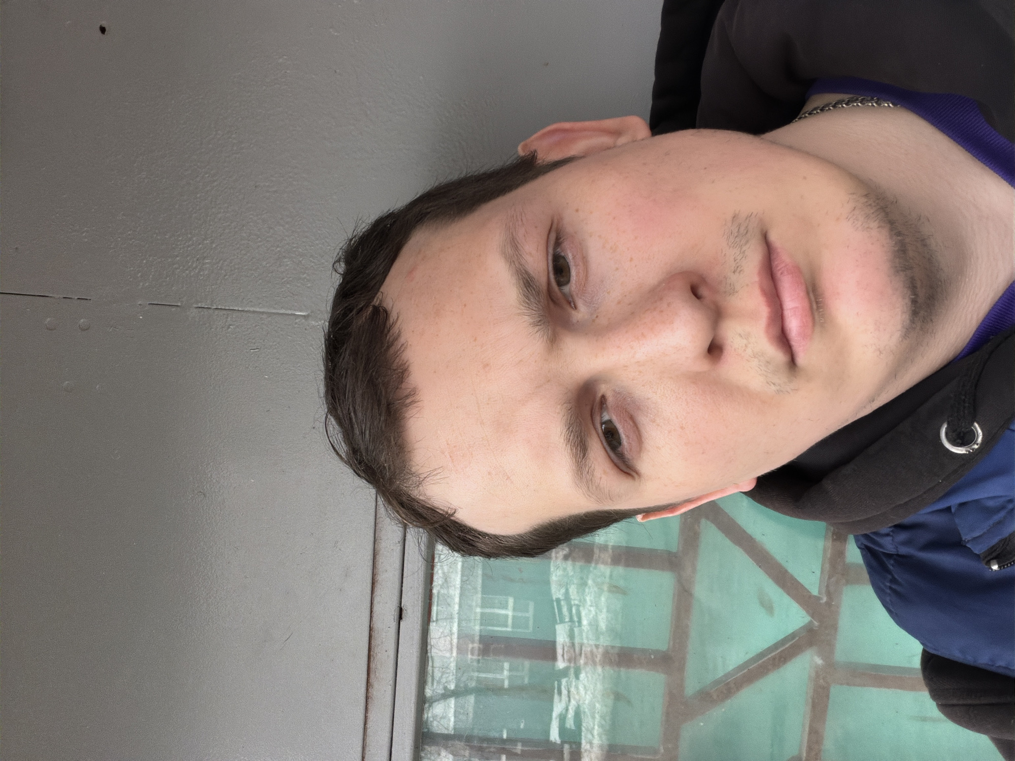 A man with short dark hair and light skin is shown from the chest up. He has brown eyes, a light beard, and a chain around his neck. He is wearing a blue and black hoodie with a purple collar. The man is standing outside with a gray wall to his left and a green window to his right. The reflection in the window shows a building and trees. The location is the city of Chaikovsky, Russia.