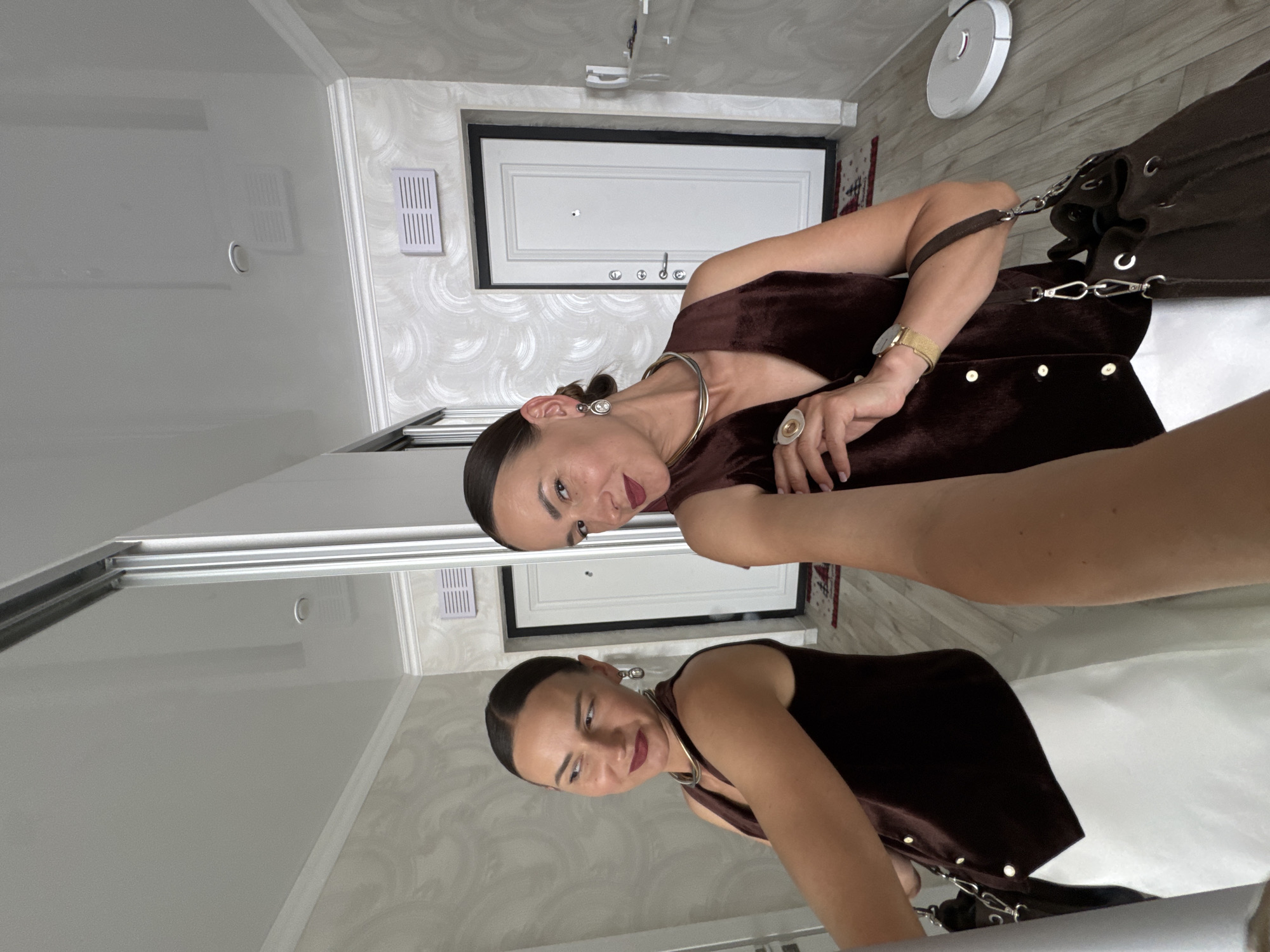 A woman is taking a selfie in a hallway. She is wearing a brown velvet vest over a white dress, a gold necklace, earrings, and a ring. She is also carrying a brown purse. The hallway has white walls with a textured pattern, white doors, and light-colored flooring. A white robotic vacuum cleaner is visible on the floor. The scene does not contain any elements that would indicate it is located in Sochi, Russia.