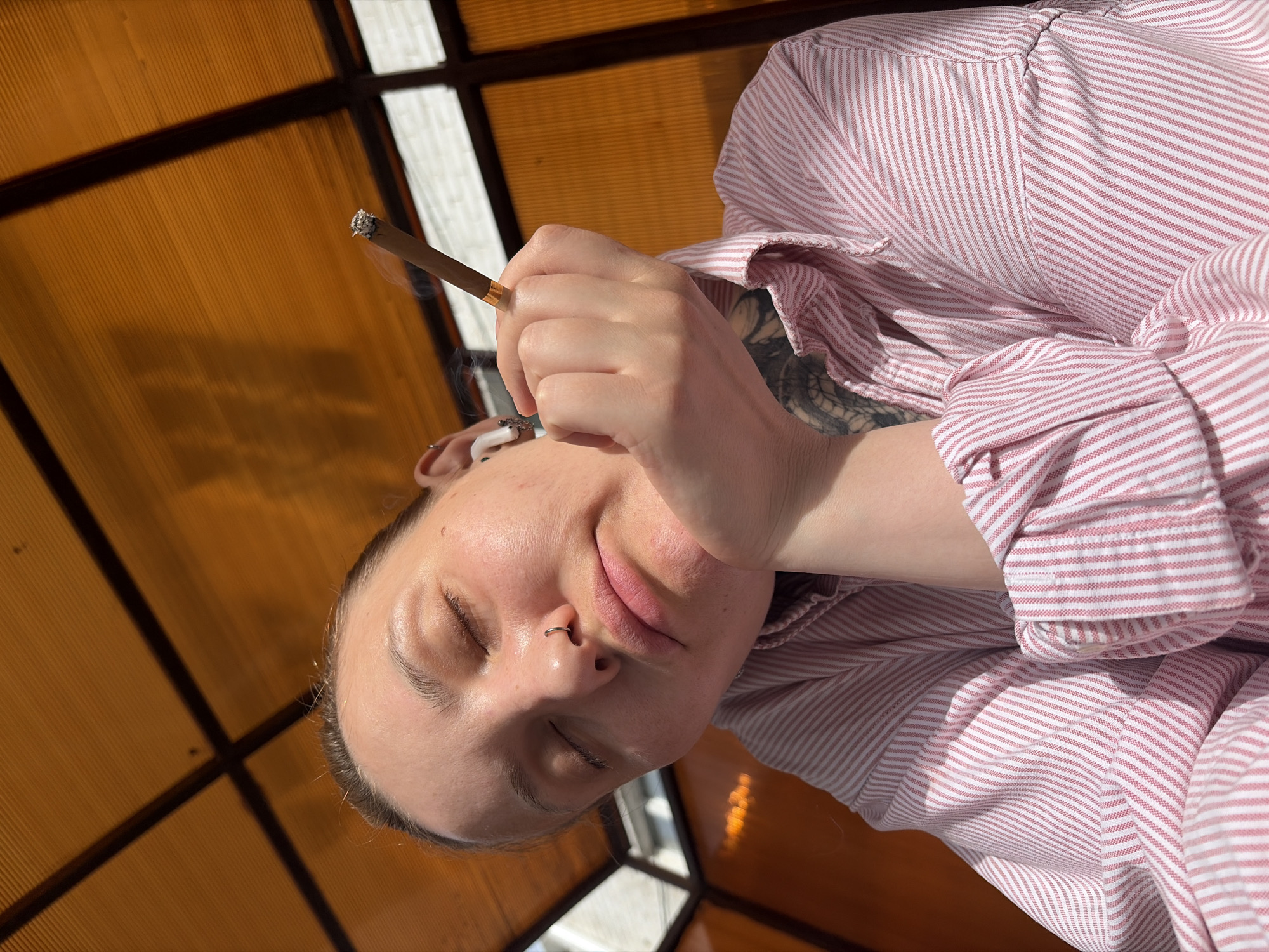 A person is holding a lit cigarette or joint. The person appears to be indoors under a translucent roof with a grid pattern. The person is wearing a pink and white striped shirt. They have a nose ring, and their eyes are closed. A tattoo is visible on their arm. No specific location is discernable, and there is no evidence to suggest it's in Chaikovsky, Russia.