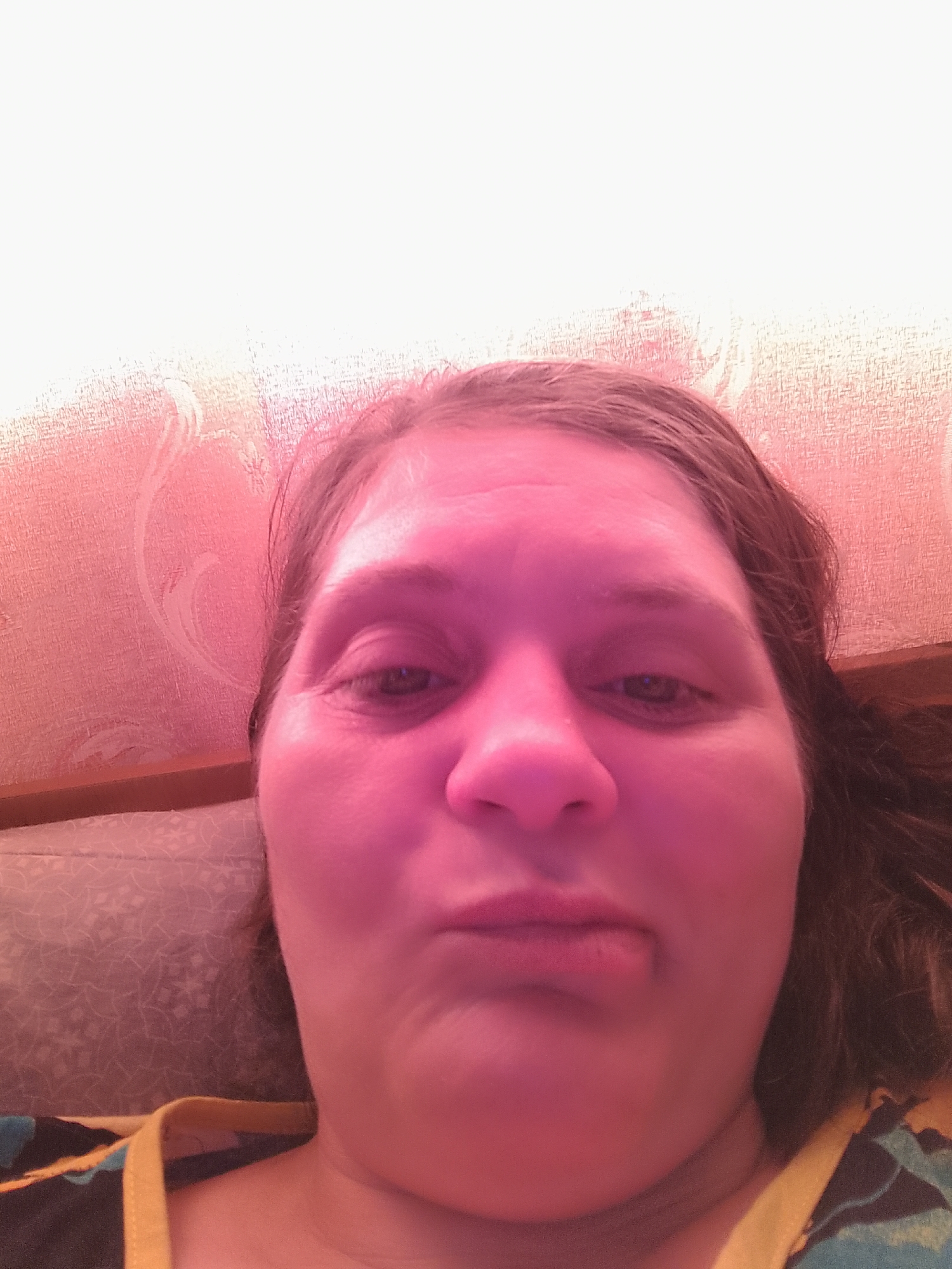 A close-up, low-angle, selfie-style photograph shows a woman's face with her mouth pursed. She has medium-toned skin and brown hair. She is wearing a yellow strap from a garment. The background features a textured, pink wall with a floral pattern, and what appears to be a patterned pillow or cushion. The lighting casts a strong pink hue over the scene, obscuring some details.