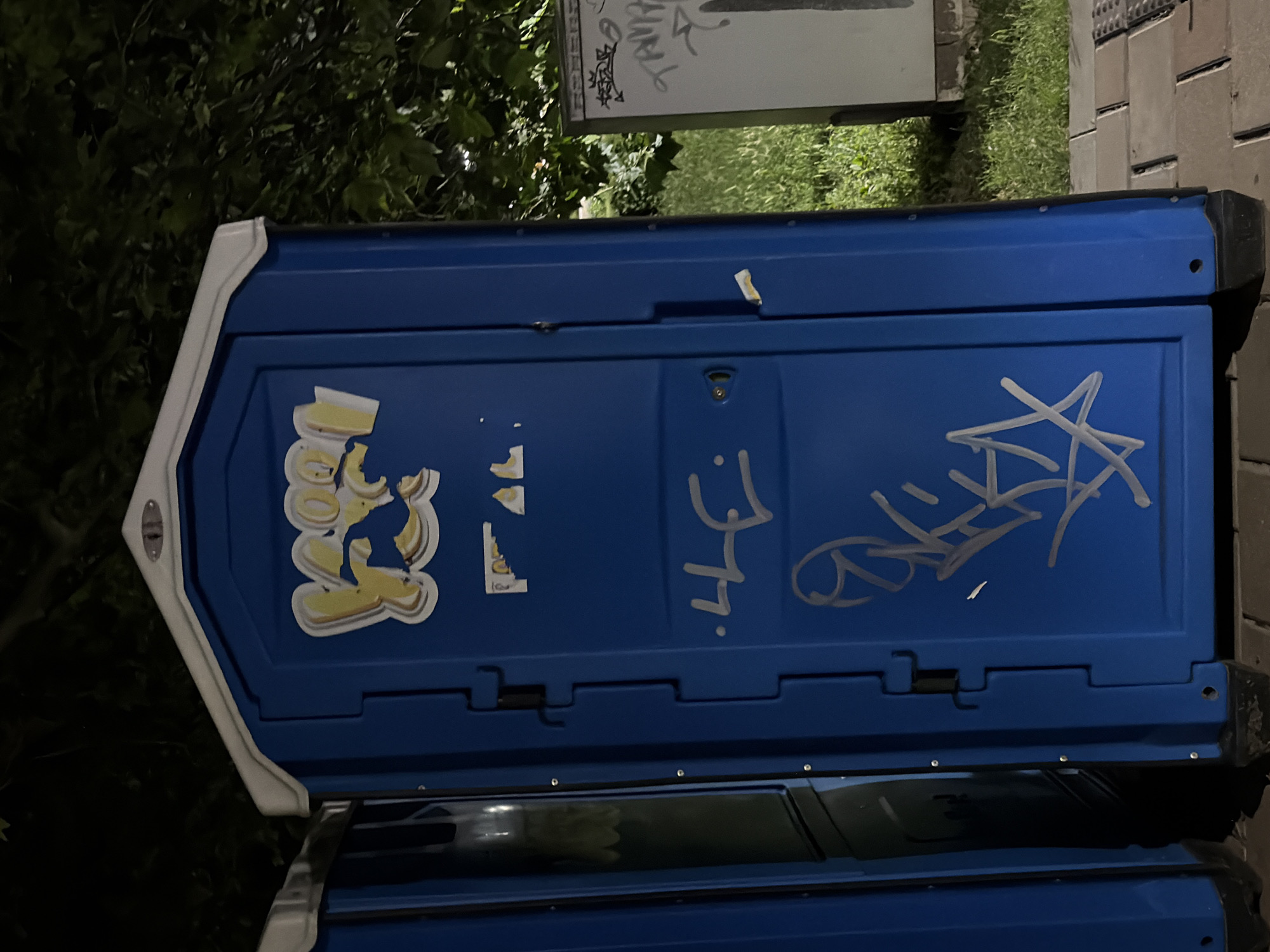 The image shows a blue portable toilet with graffiti on it. The portable toilet is located outdoors, with trees and grass in the background. There are other portable toilets to the left and a white structure with graffiti to the right. The ground is paved with bricks. The graffiti on the blue portable toilet appears to be in yellow and white. The yellow graffiti on the top of the door has been partially obscured by white markings. There are also white graffiti tags further down the door.