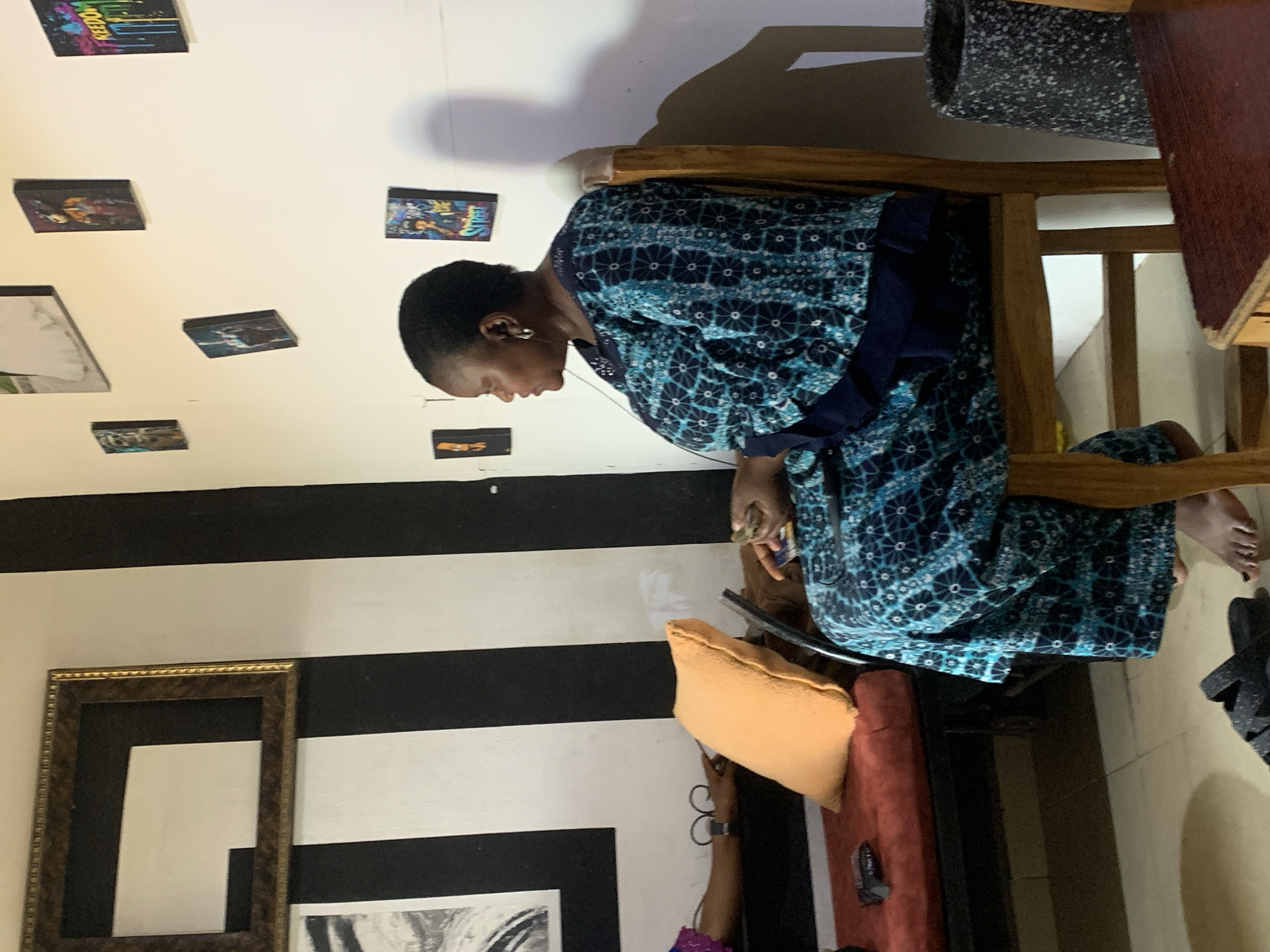 A woman sits on a wooden chair, wearing a blue patterned robe and listening to earbuds. She appears to be using a smartphone or tablet. The room has a wall with black and white horizontal stripes and framed artwork. The location is suggested to be in Jos, Nigeria. Additional details include an orange pillow, a glimpse of a seated person with purple fabric, and what appears to be a rug or padding on top of the chair.