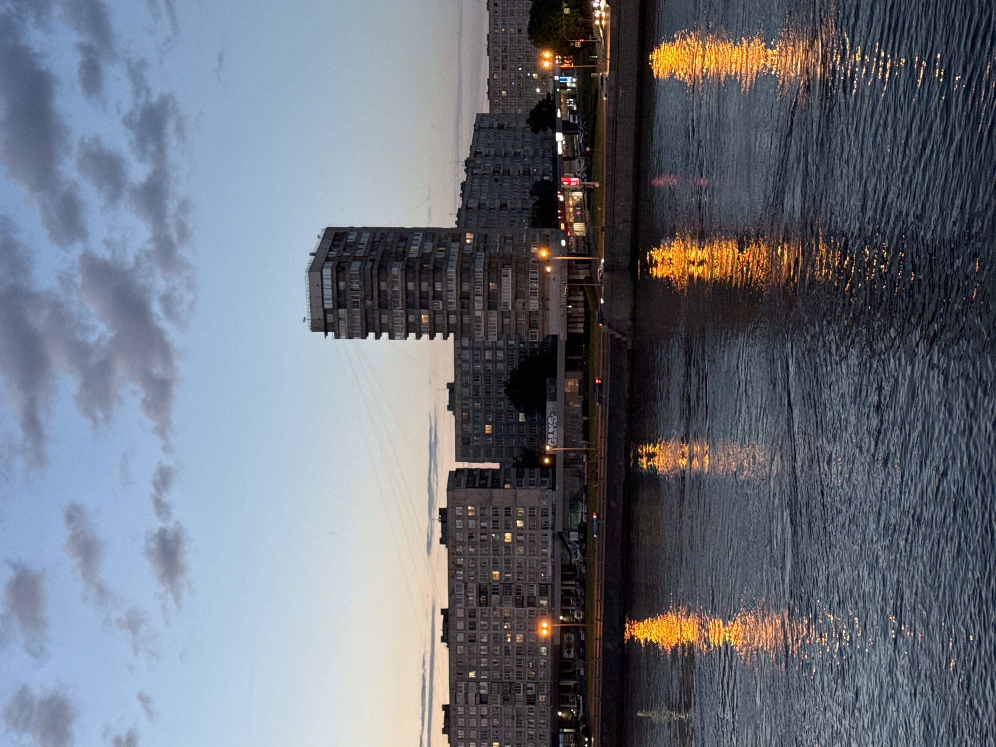 This image shows a city skyline at dusk, with the Neva River in the foreground. Several large, multi-story residential buildings line the riverbank, some with windows illuminated by interior lights. A tall, slender skyscraper stands out among the apartment blocks. The sky is a gradient of pale blue to orange, with scattered clouds. The river reflects the lights from the buildings and streetlights, creating shimmering streaks of orange and yellow on its dark surface. A concrete embankment separates the river from the buildings, and a road with vehicles is visible in front of the buildings. The scene appears to be in Kudrovo, Russia, a suburb of Saint Petersburg, known for its modern residential developments. There are no identifiable people or specific actions depicted. The overall impression is one of urban life at twilight, with the natural element of the river contrasting with the man-made structures.