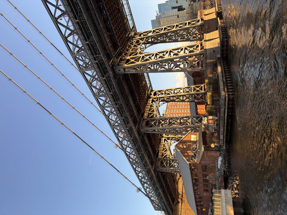 The image captures a detailed view of the Manhattan Bridge's underside and base structure from the East River in New York City, United States. The photograph is taken from a low angle, showcasing the massive steel-truss architecture of the bridge, characterized by intricate gray lattice beams and robust support pillars. Multiple suspension cables extend diagonally upwards into a clear blue sky, which contains a faint white aircraft contrail in the upper left quadrant.
Below the bridge's complex metallic framework, a long, low-rise brick building with a pitched roof is situated near the water on the left side, displaying several windows and graffiti on its facade and roof. To the right and further under the bridge, additional smaller brick structures and platforms supported by concrete piles are visible, some also bearing graffiti. A rocky shoreline segment is present near the leftmost brick building.
The East River occupies the lower portion of the frame, showing ripples on its surface and reflecting warm golden sunlight, suggesting either early morning or late afternoon. In the background, a multi-story reddish-orange brick building stands behind the bridge's supports, while modern high-rise buildings with reflective surfaces are discernible further in the distance on the right, across the river. No human figures are present in the scene.