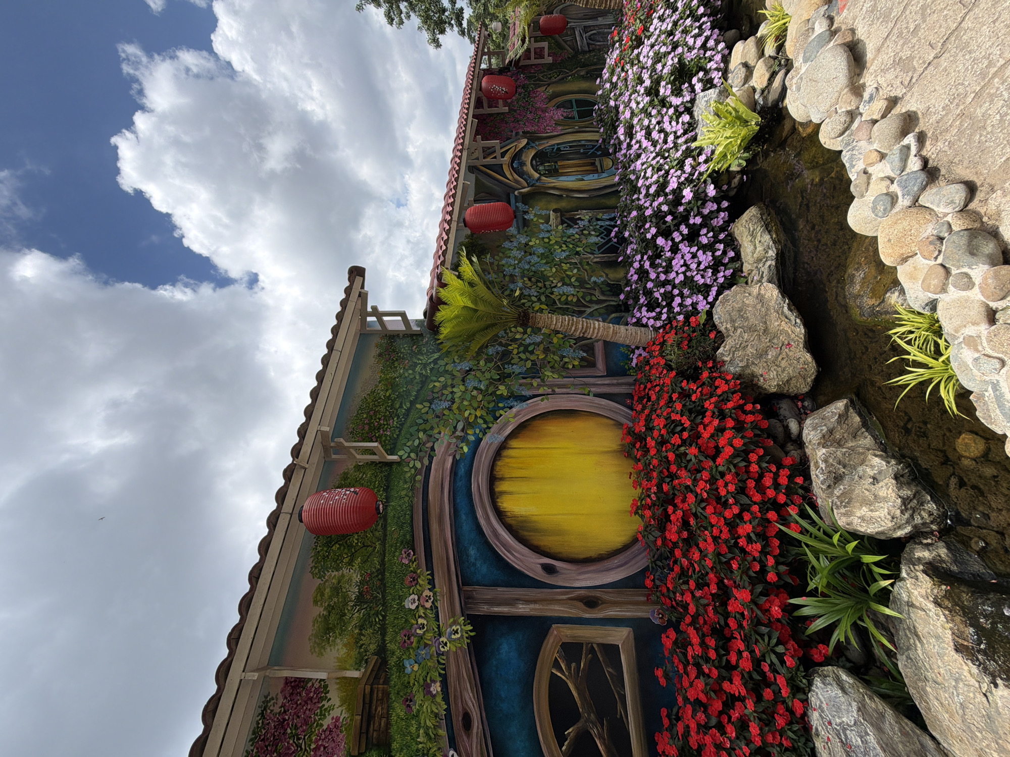 The image depicts a whimsical, fairy-tale-inspired building facade, likely a tourist attraction in Da Lat, Vietnam, given the style reminiscent of " Hobbit homes" popular in the area. The structure is painted with vibrant murals, creating the illusion of round doors and windows set into a grassy hillside. Red and pink flowers are planted profusely in front of the building, bordering a small pond with smooth, rounded stones. Several red paper lanterns hang from the eaves of the building, adding to the decorative theme. The sky above is a mix of blue and white clouds.
