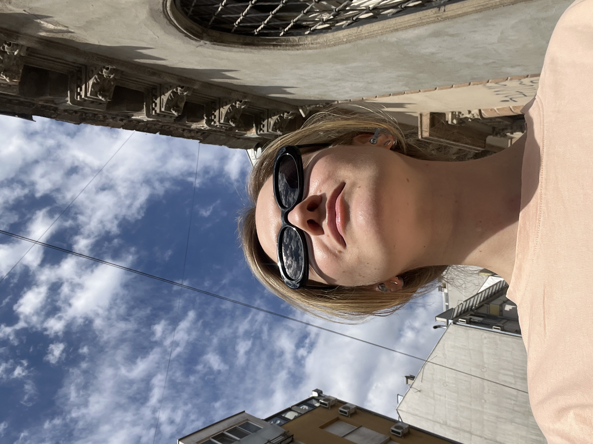 A low-angle, outdoor shot shows a fair-skinned young woman wearing sunglasses and a light pink top. Buildings and a sky filled with white clouds form the background. Architectural details of a building, including an ornate window, are visible above her head. In the lower part of the frame, more modern buildings are present. Electrical wires stretch across the sky. It appears to be a sunny day in Novi Sad, Serbia.
