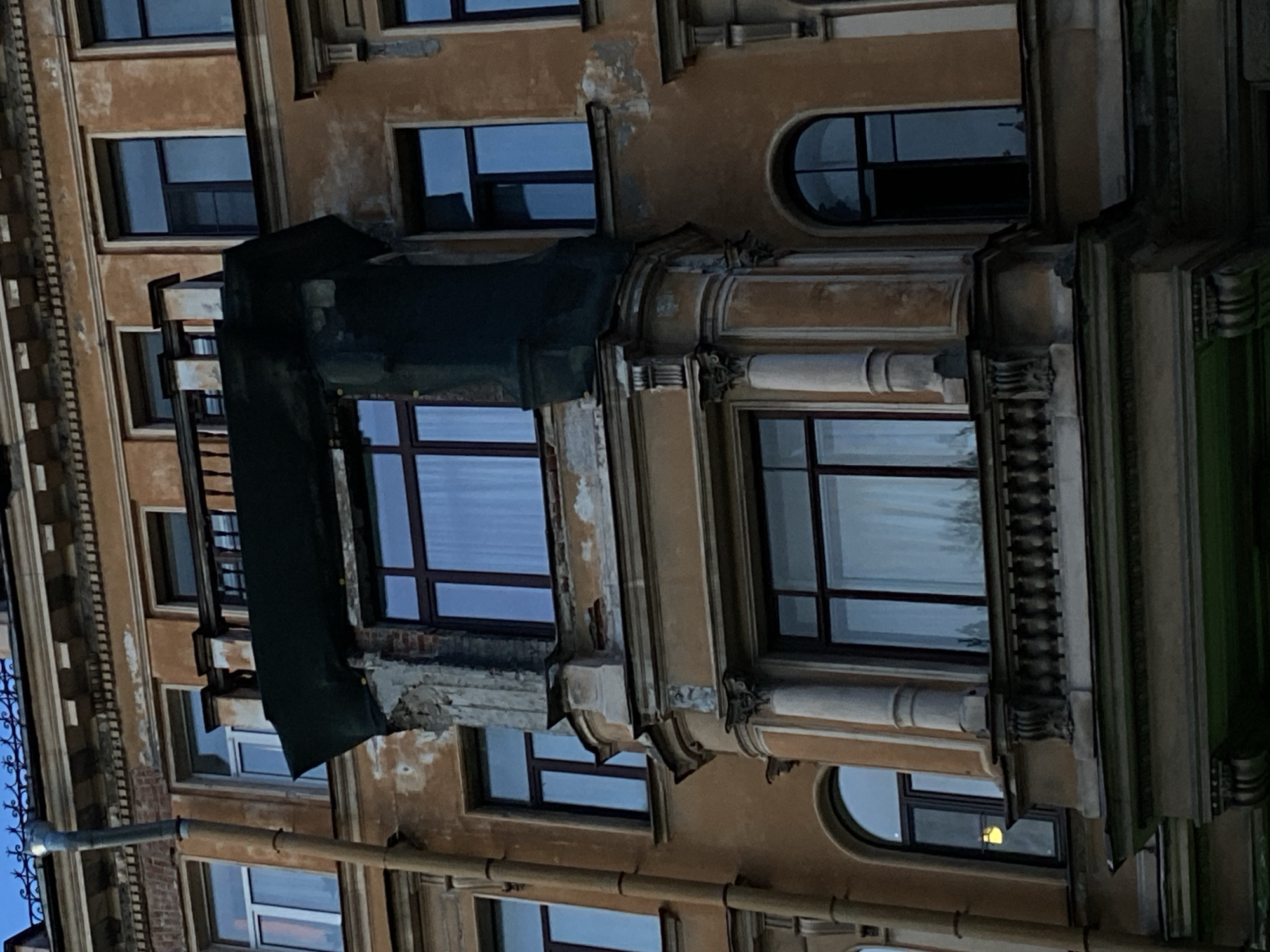 An upward angle shot captures a section of a building in St. Petersburg, Russia. The building is characterized by a faded yellow-brown facade, detailed window frames, and varied window shapes (rectangular, rounded). A dark green tarp obscures part of one window, and some facade sections show decay. The scene is primarily lit by ambient light, indicating it is night or dusk.