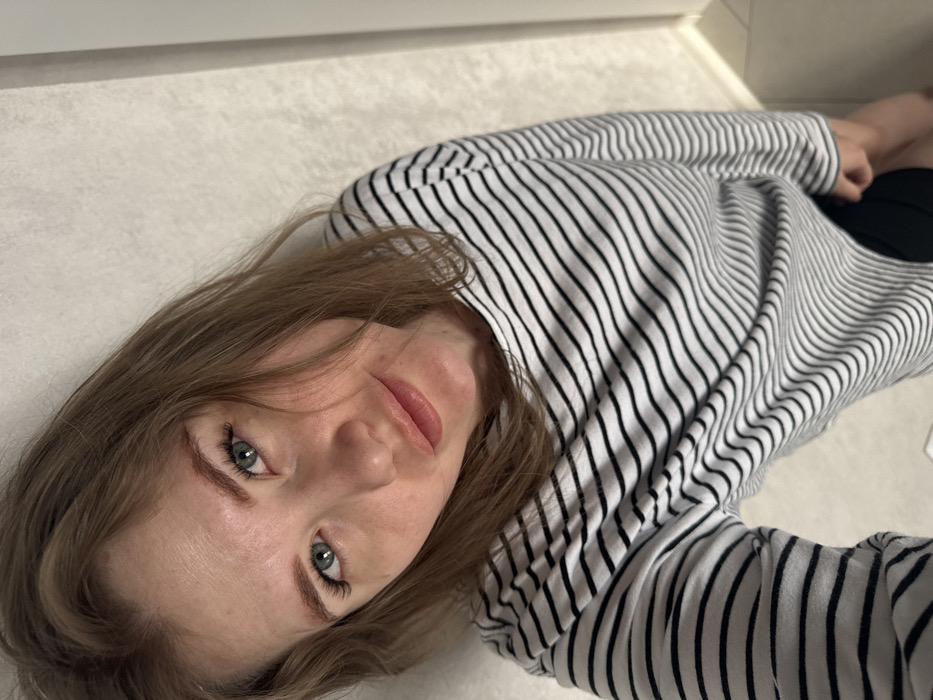 The image presents an adult female in an indoor setting within Gornyi (Dvurechenskoe S/P), Russia. The subject has light brown hair, blue-green eyes with defined eyelashes, and light-colored, shaped eyebrows. Her lips are full and tinted light pink. She is wearing a long-sleeved shirt with horizontal black and white stripes, with a dark lower garment partially visible at the bottom right. The individual is looking directly into the camera, which appears to be held at a slightly low angle. The background consists of a plain, light-colored wall, possibly textured, with a vertical line or corner visible on the right side.